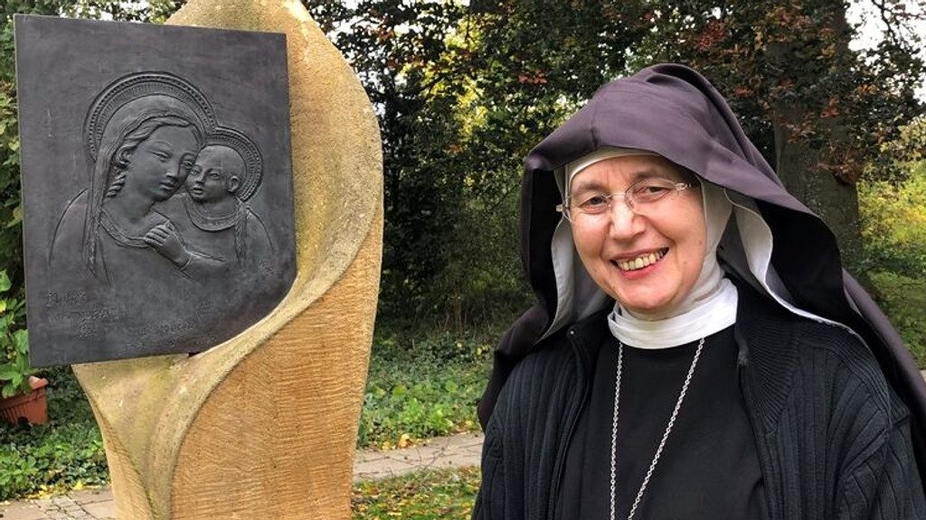 Eine Ordensfrau im schwarzen Habit steht in einem Garen neben einer Holzskulptur.