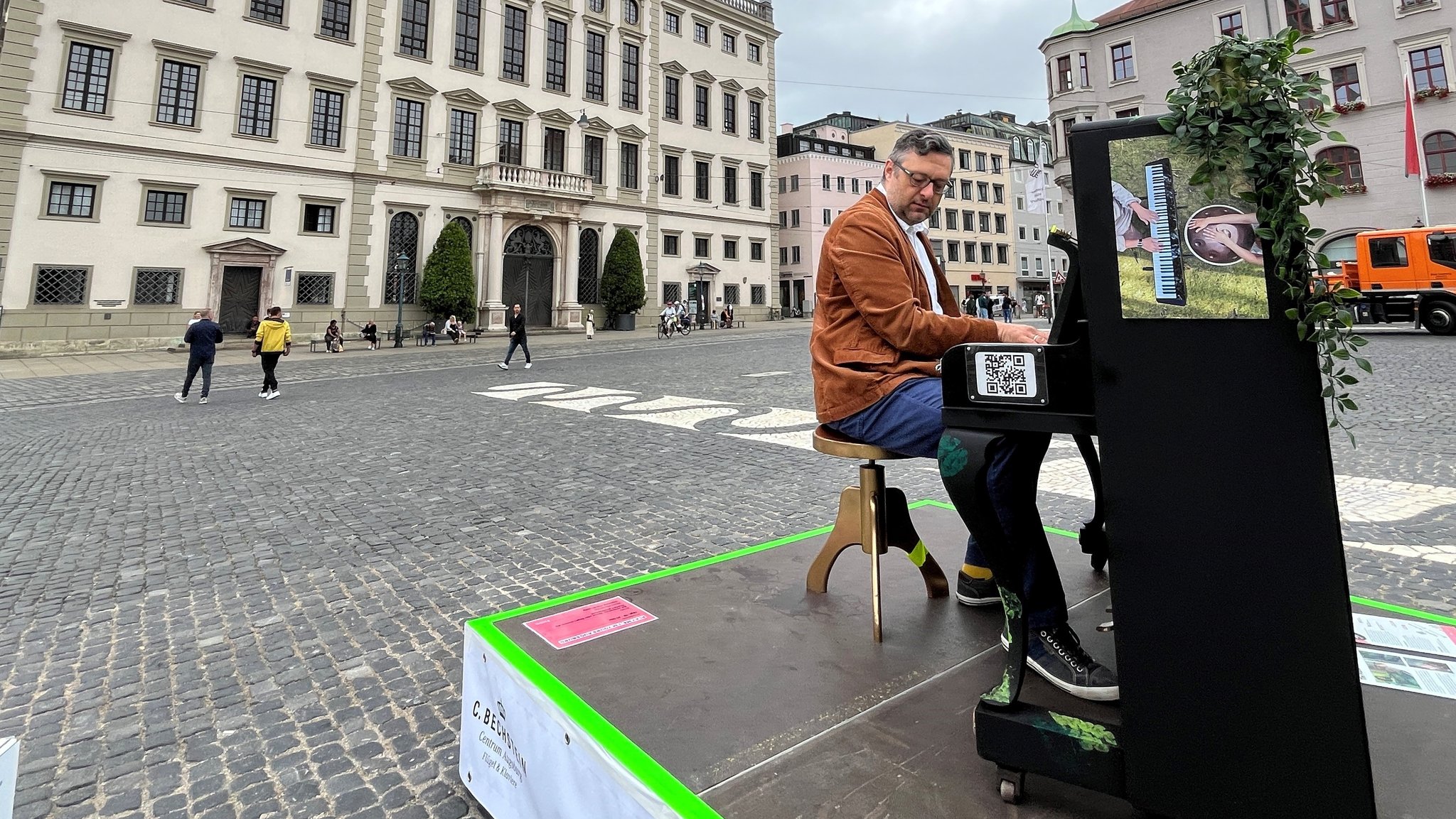 BR-Mitarbeiter Jochen Eichner sitzt in weißem Hemd und brauner Jacke an einem Klavier in der Mitte des Rathausplatzes und spielt es mit gefühlvollem Gesichtsausdruck. Passanten laufen vorbei. 