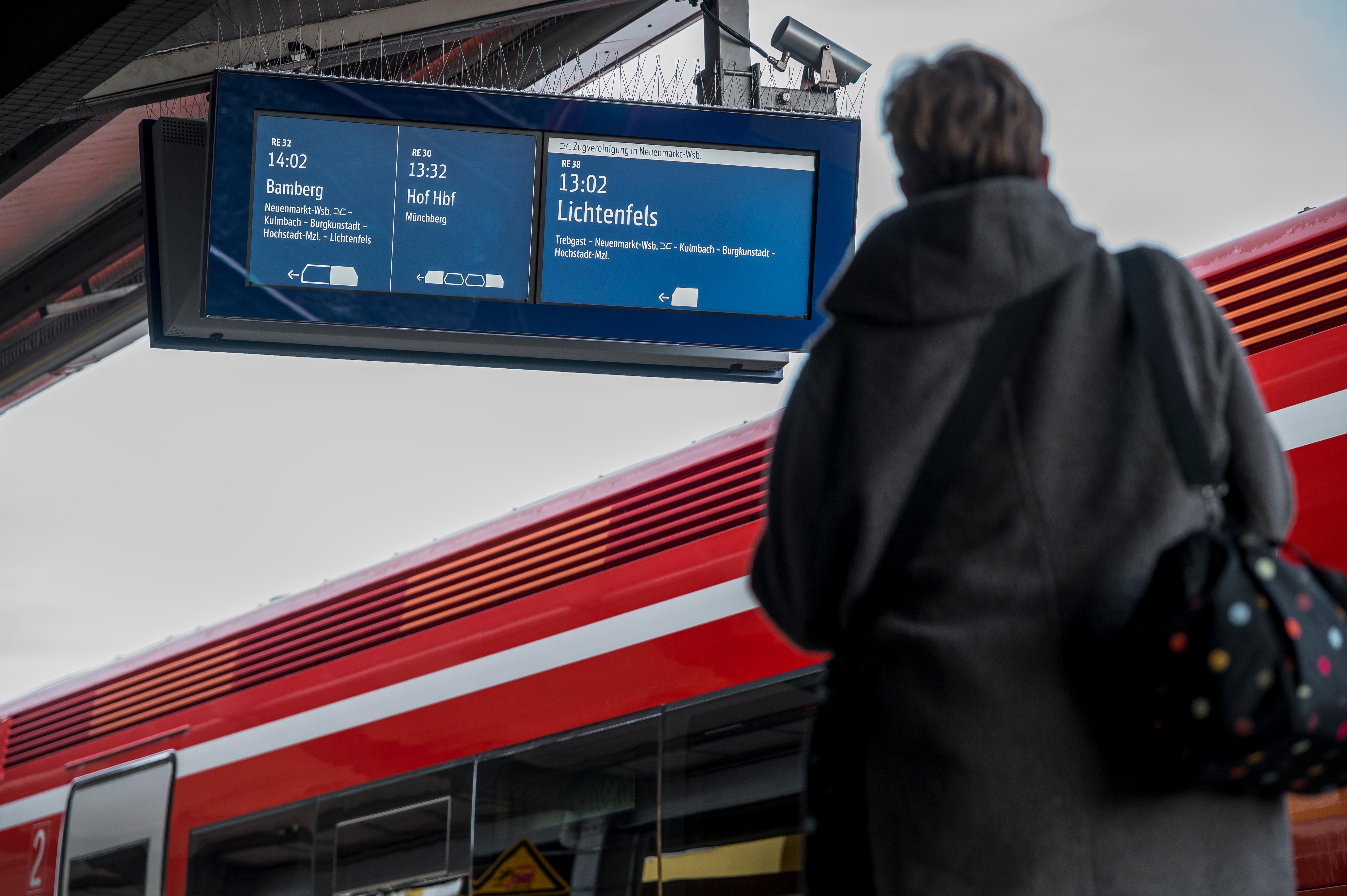 Eine Frau steht am Bahngleis vor einem Zug. Auf der Anzeigetafel sind Verbindungen nach Bamberg, Hof und Lichtenfels angegeben.