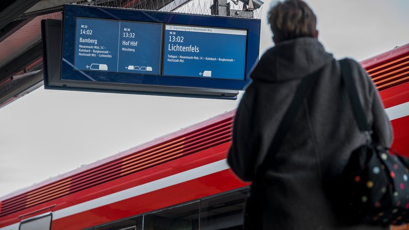 Eine Frau steht am Bahngleis vor einem Zug. Auf der Anzeigetafel sind Verbindungen nach Bamberg, Hof und Lichtenfels angegeben.