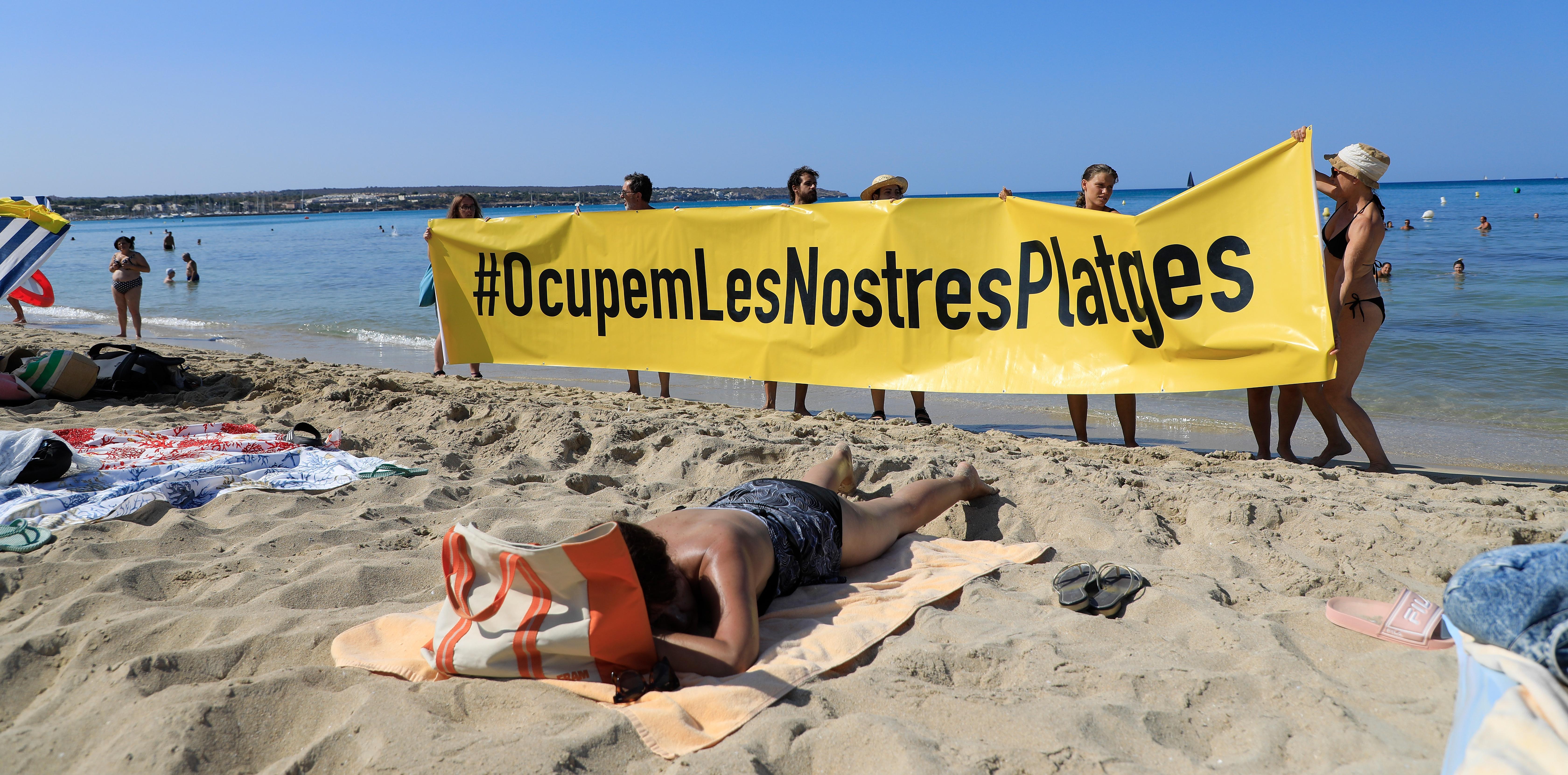 Menschen halten an einem Strand auf Mallorca ein Banner "#Sie besetzen unsere Strände" aus Protest gegen den Massentourismus auf der Balearen-Insel hoch.