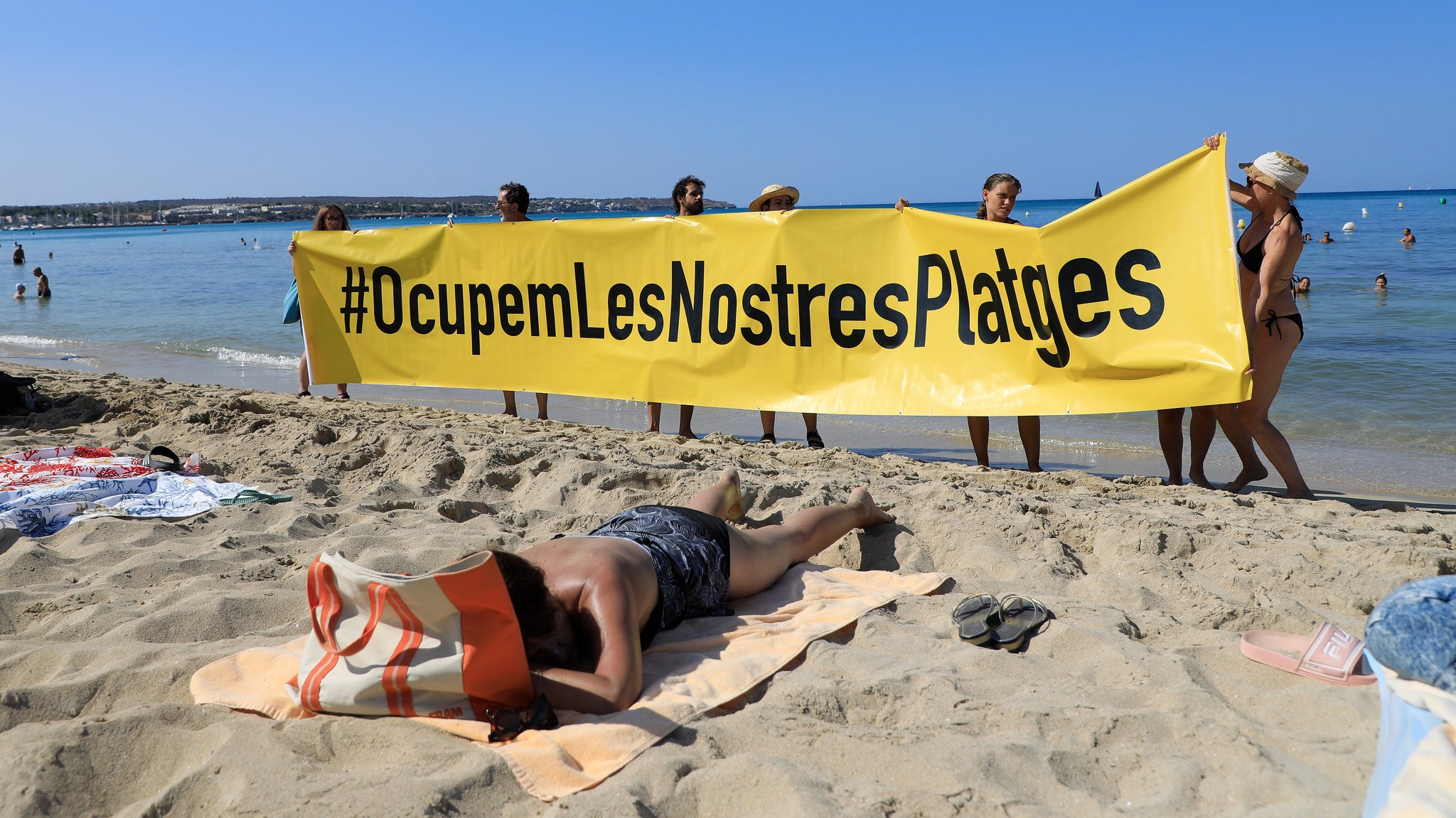 Menschen halten an einem Strand auf Mallorca ein Banner "#Sie besetzen unsere Strände" aus Protest gegen den Massentourismus auf der Balearen-Insel hoch.