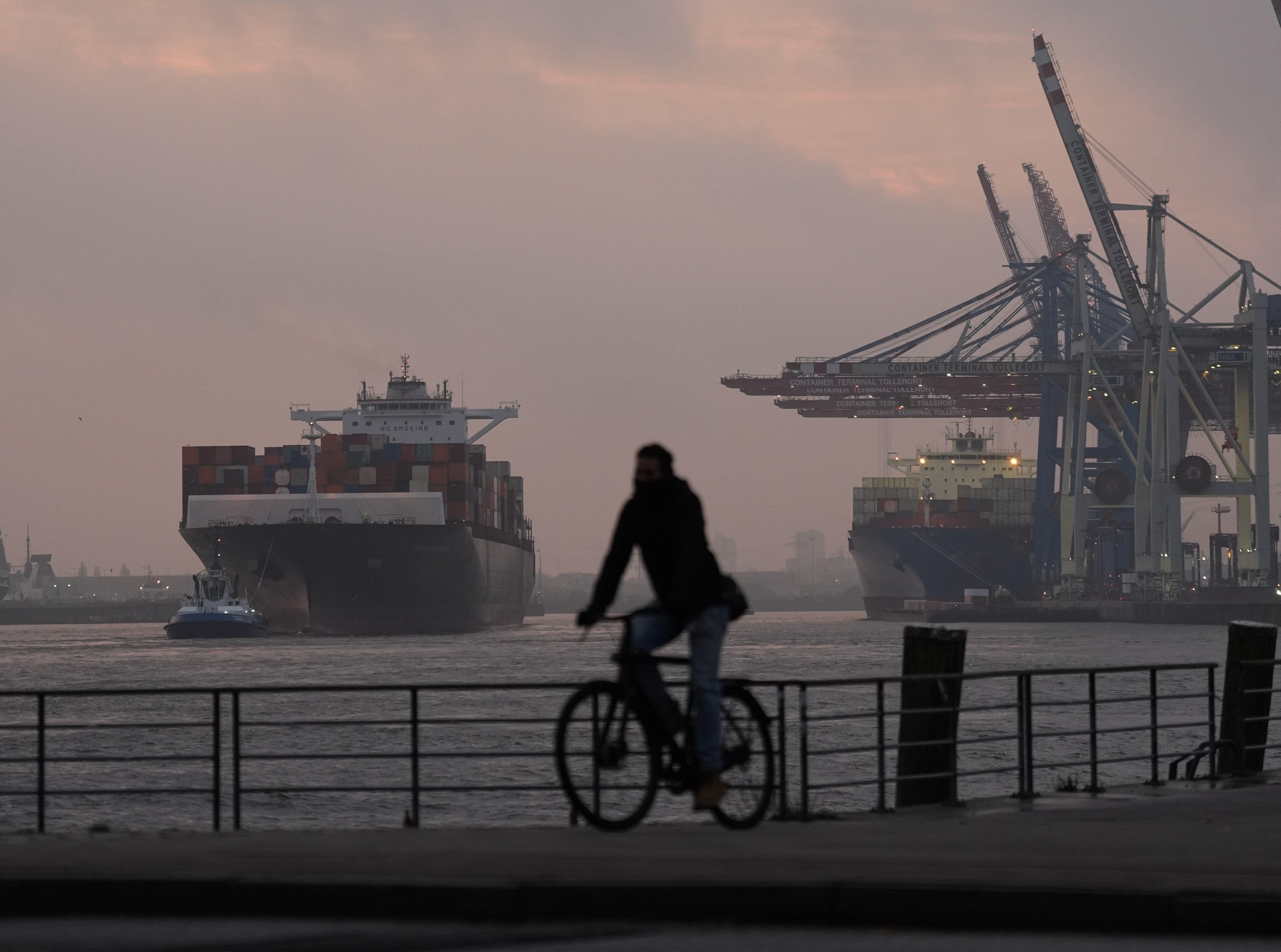 Zu sehen ist ein Containerschiff im Hamburger Hafen. Ein Fahrradfahrer fährt entlang der Promenade.