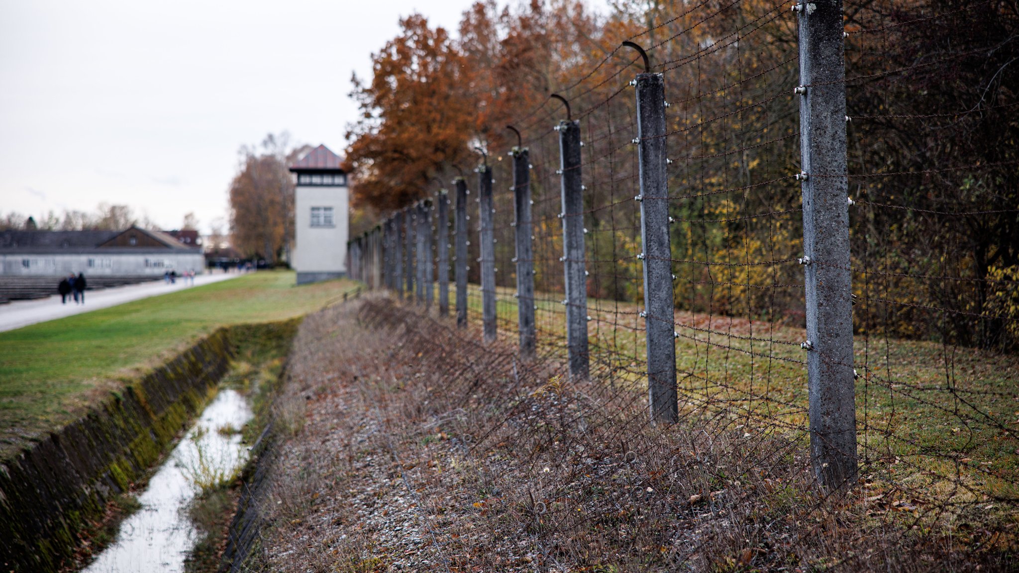 Zaun mit Stacheldraht und Wachturm an einer Kontrollstraße auf dem Gelände der KZ-Gedenkstätte Dachau.