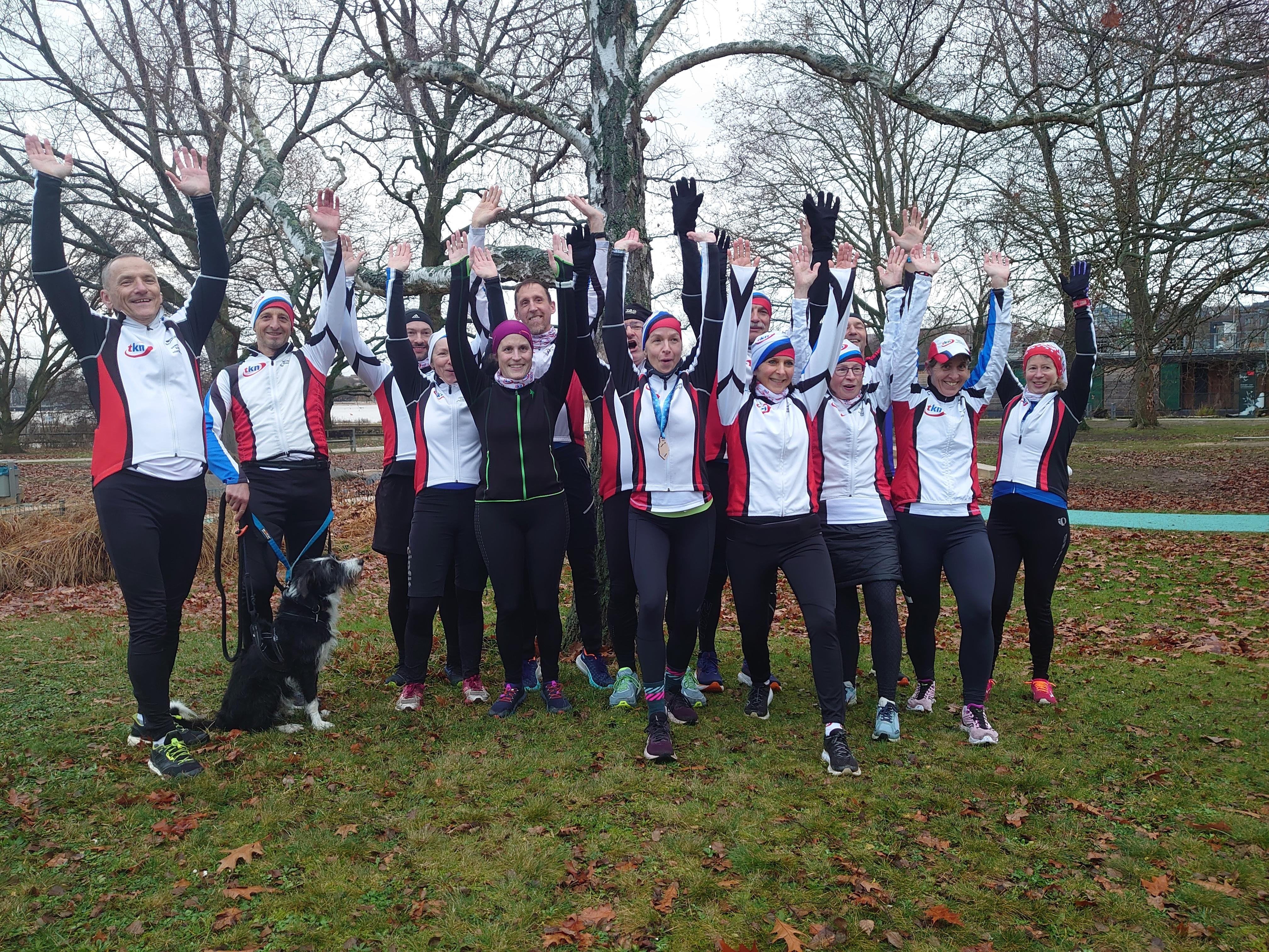 Vierzehn Läuferinnen und Läufer vom Verein Team Klinikum Nürnberg stehen für ein Gruppenfoto zusammen und heben die Arme in die Luft.