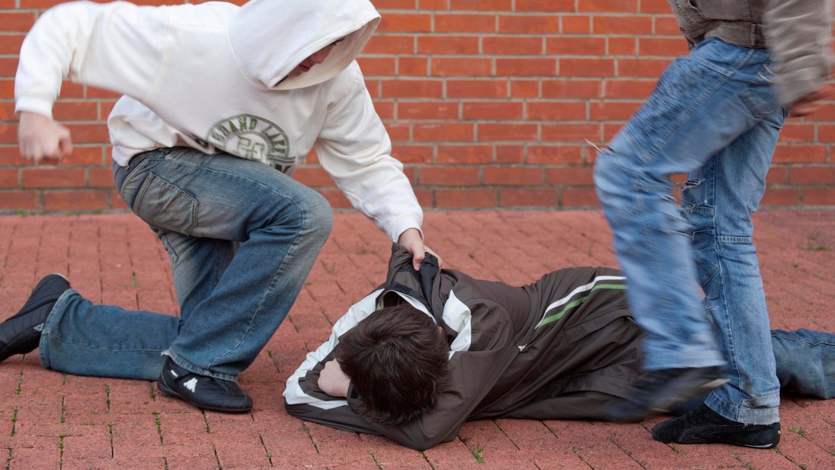 Ein am Boden liegender wehrloser Junge wird von zwei anderen brutal festgehalten und getreten (gestellte Szene).