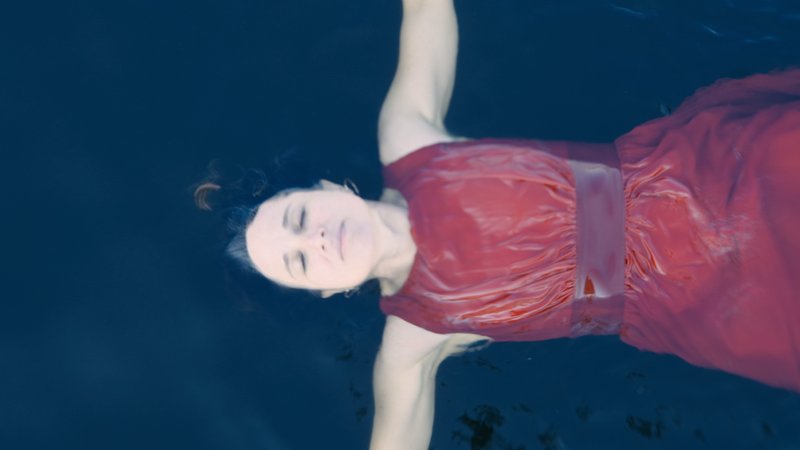 Eine Frau in rotem Kleid schwimmt gleitend in einem Pool, Szene aus "Anima – Die Kleider meines Vaters".