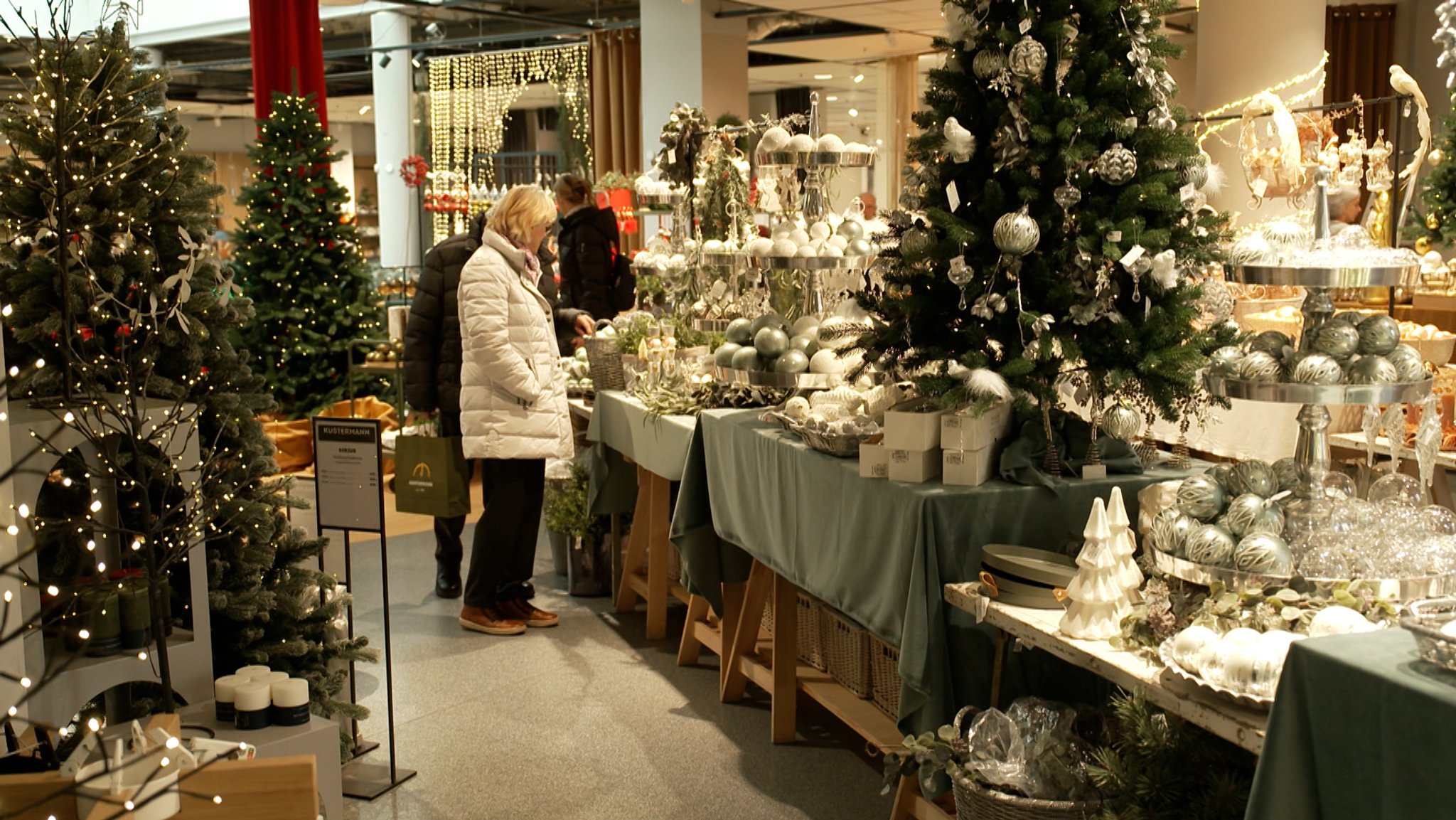 Eine Frau und ein Mann in Winterjacken stehen zwischen Tischen mit weißer und silberfarbener Advents-Deko in einem Geschäft. Der Advent ist für die Einzelhandelsgeschäfte die Zeit mit den höchsten Umsätzen - der bayerische Handelsverband hofft auf gute Geschäfte.