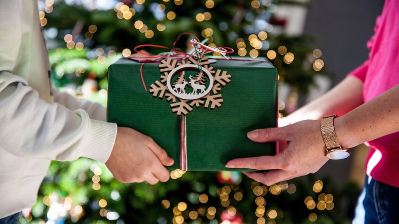 Zwei Personen reichen sich ein weihnachtlich verpacktes Geschenk. im Hintergrund sieht man den Christbaum, Weihnachtsbaum stehen. 