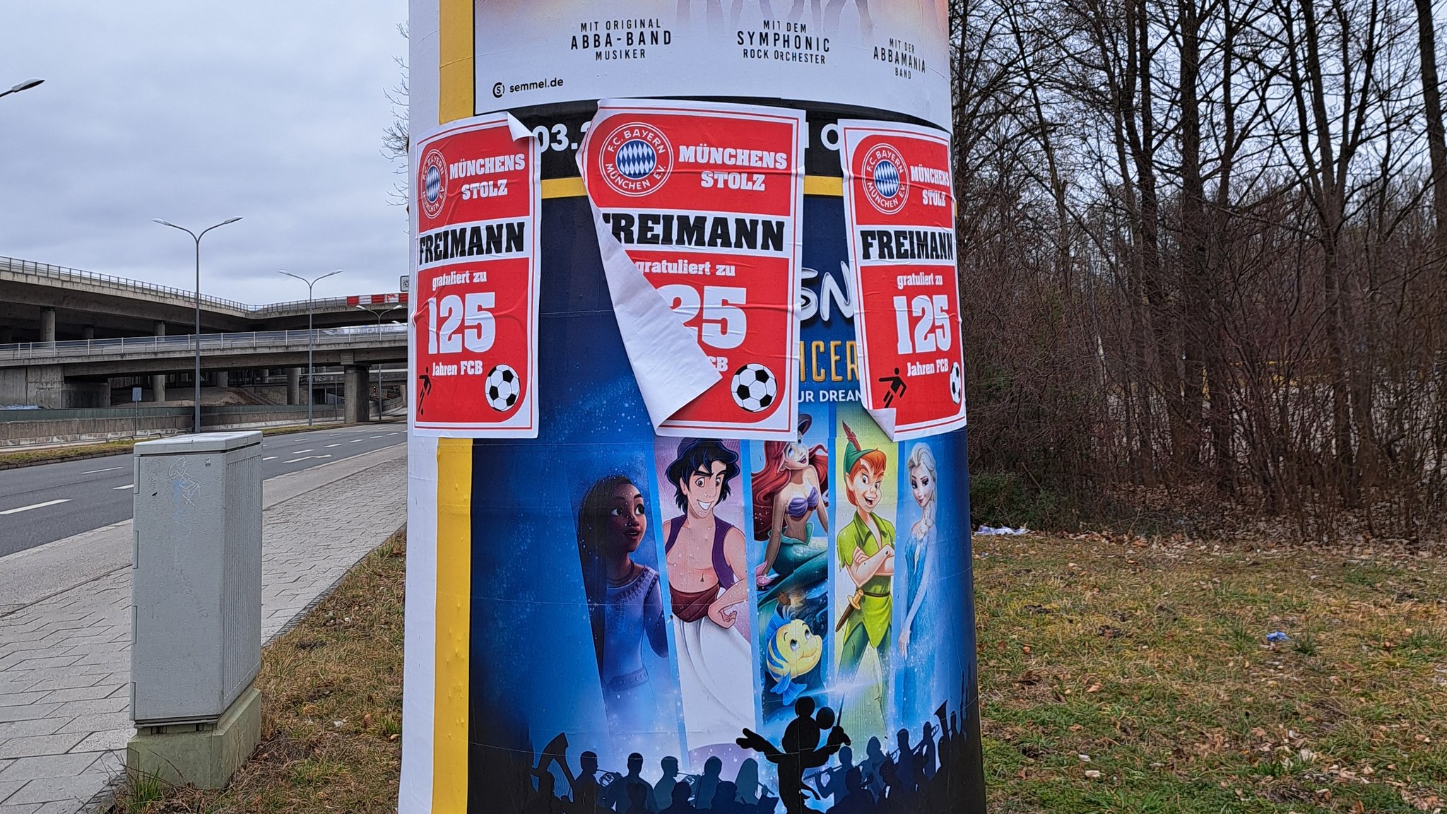 Auf einer Litfasssäule an einem Straßenrand in München kleben DIN-A-4-große Aufkleber mit der Aufschrift "Freimann gratuliert zu 125 Jahren FCB" über den Plakaten zu einer Veranstaltung.