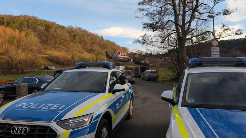 Einsatzfahrzeuge der Polizei stehen auf einer Straße. Dahinter sind weitere Pkw zu sehen. Im Hintergrund ist eine Straße zu sehen die nach Altschauerberg führt. | Bild: BR/Nicolas Eberlein Einsatzfahrzeuge der Polizei stehen auf einer Straße. Dahinter sind weitere Pkw zu sehen. Im Hintergrund ist eine Straße zu sehen die nach Altschauerberg führt.