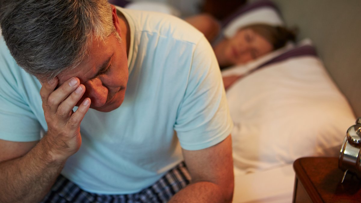 Ein älterer Mann sitzt nachts am Bett und hält sich eine Hand an den Kopf. Er kann nicht schlafen, weil er sich unwohl fühlt.