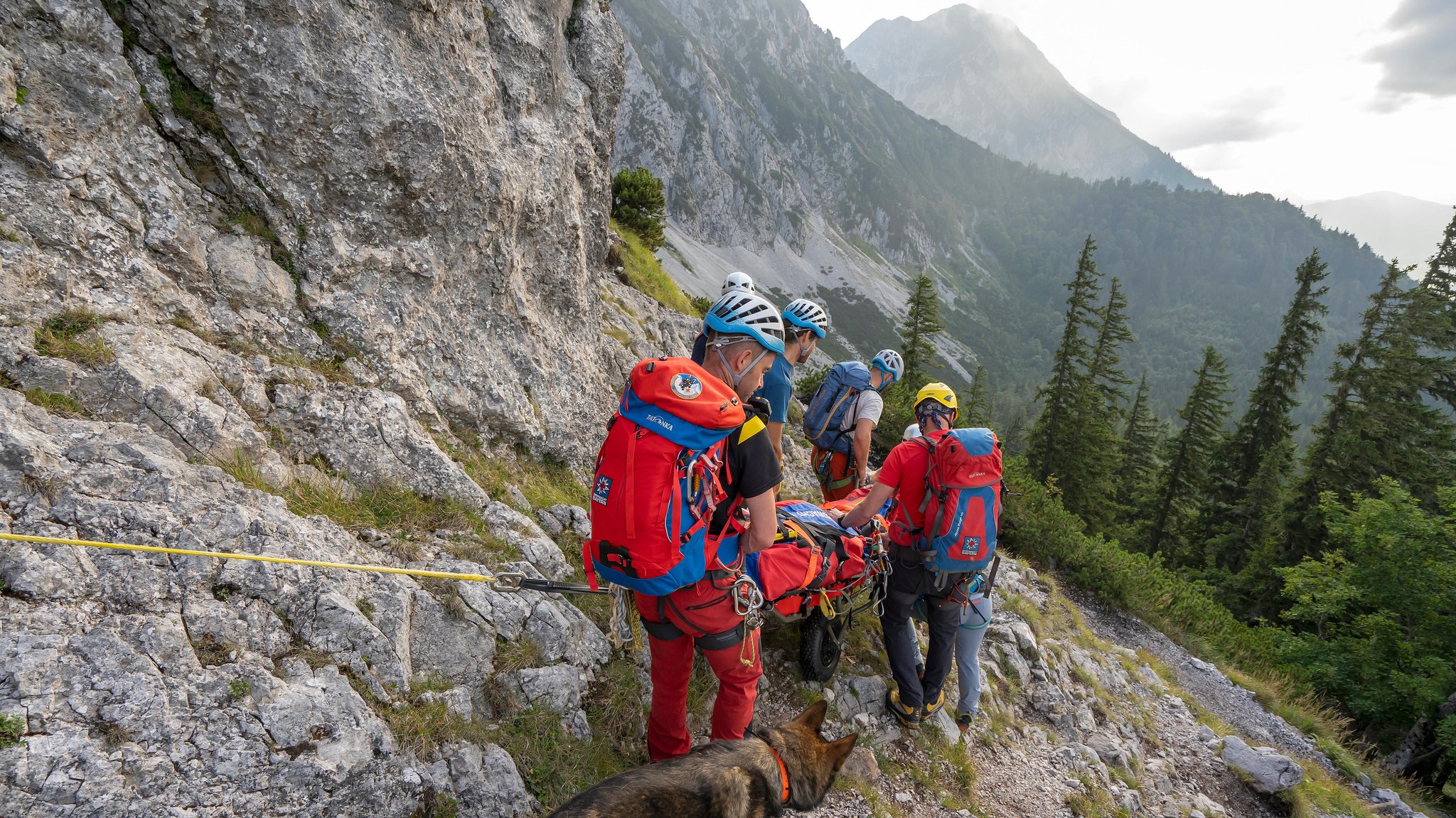 Bergwachtler transportieren einen Verletzten in der Trage über felsiges Gelände. In der Ferne sind Gipfel zu sehen. Das Bild stammt von einer Sommerübung am Hochstaufen.