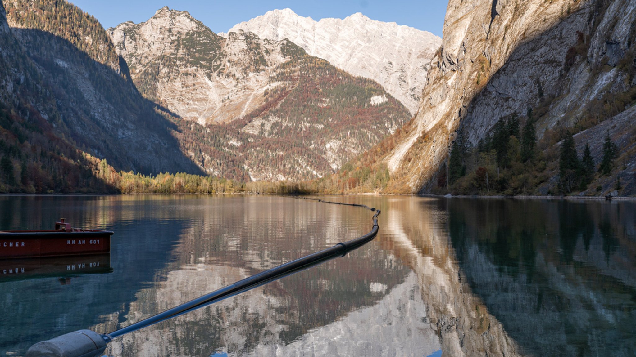 Ein dunkles Rohr zieht sich über die Wasseroberfläche des Königssees, im Hintergrund Bergmassiv