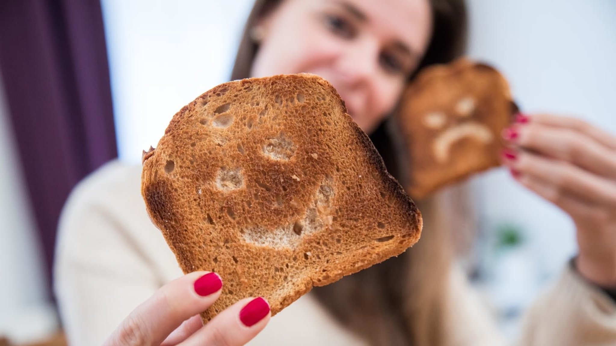 Eine junge Frau hält zwei Scheiben Toastbrot mit eingeritzten Gesichtern in den Händen.