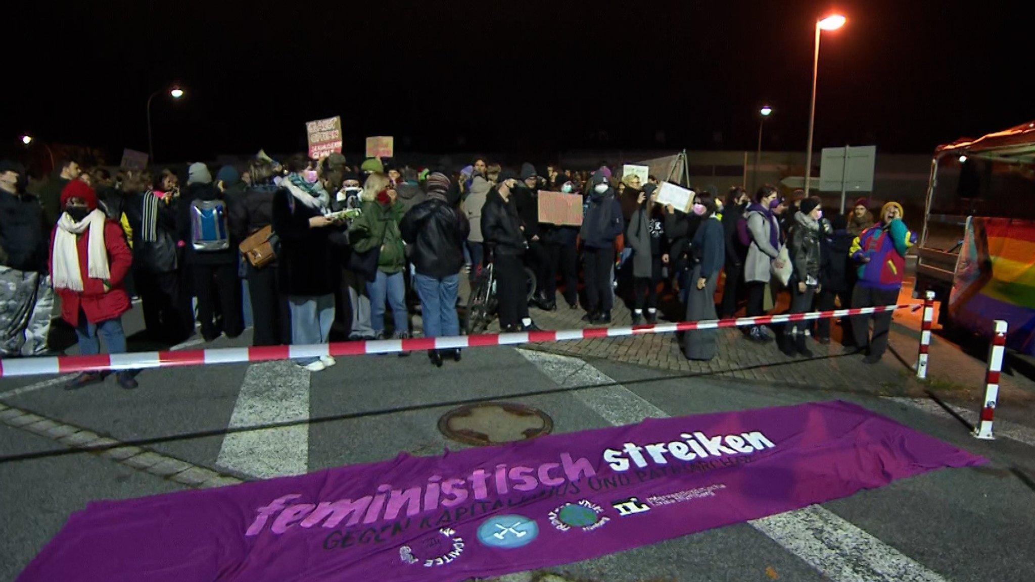 Hinter einem rot-weißen Absperrband stehen Menschen und halten Plakate in die Höhe. Auf der Straße liegt ein Transparent mit der Aufschrift "feministisch streiken". 