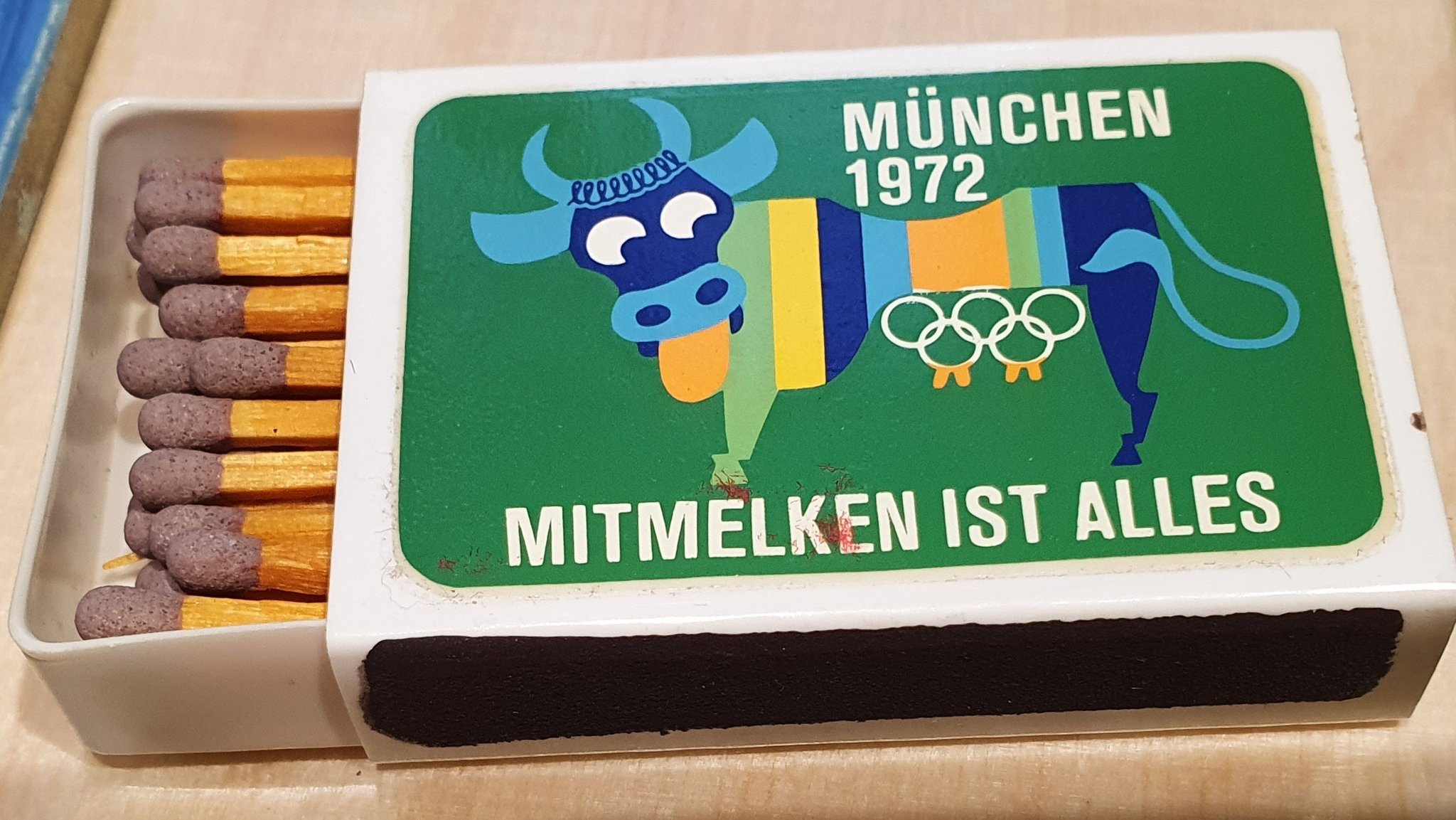 Zündholzschachtel aus dem Olympia-Jahr 1972 mit buntgestreifter Kuh und Inschrift "Mitmelken ist alles"