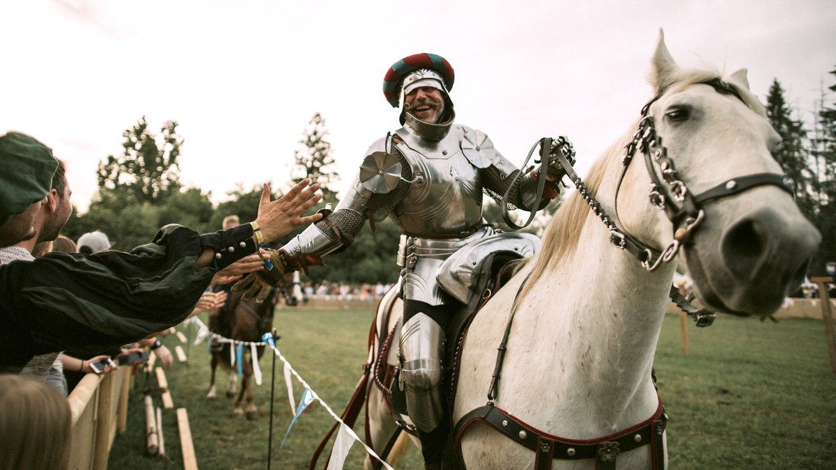 Bild von 2019: Ein Mann mit Schnauzer in Ritterrüstung auf einem weißen Pferd klatscht mit einem Mann in mittelalterlicher Kleidung ein.