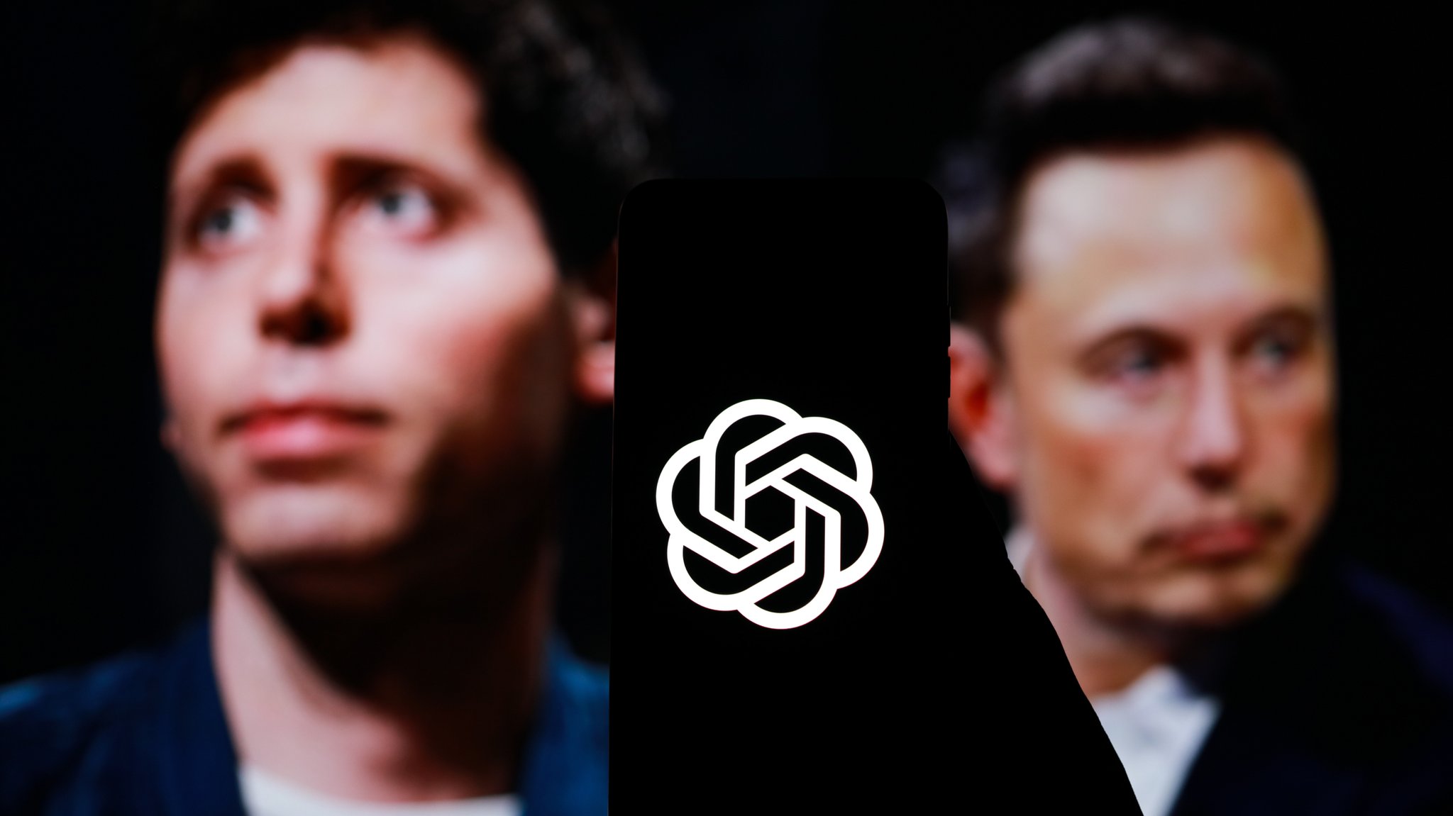 Sam Altman und Elon Musk verschwommen zu sehen hinter einem OpenAI-Logo. | Bild: picture alliance / ZUMAPRESS.com | Ahmed Fesal Bayaa Sam Altman und Elon Musk verschwommen zu sehen hinter einem OpenAI-Logo.