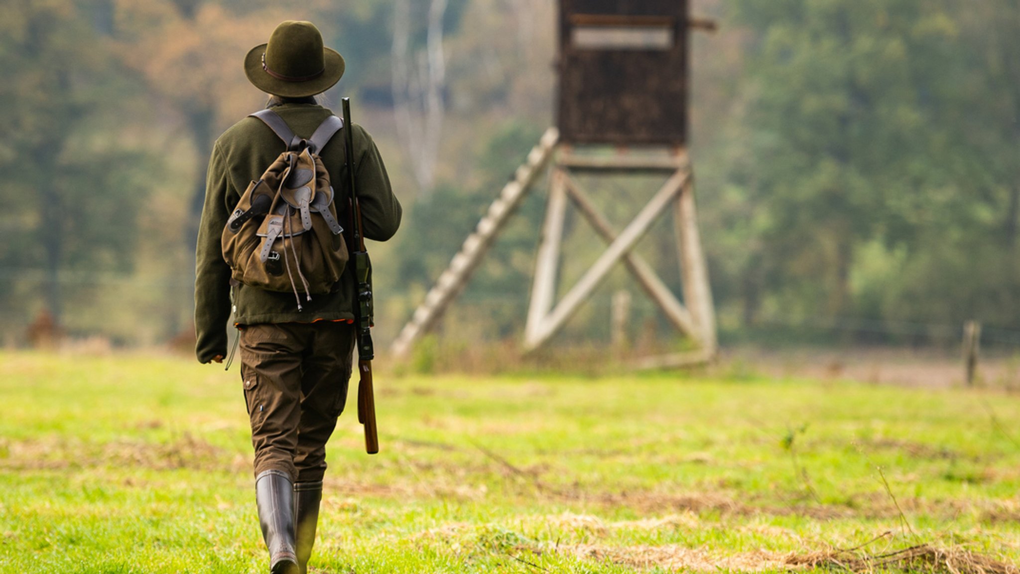 Eine Frau in olivgrüner Kleidung, Gummistiefeln, mit Hut, Gewehr und Rucksack ist von hinten zu sehen, wie sie auf einen im Hintergrund stehenden Hochsitz zuläuft.