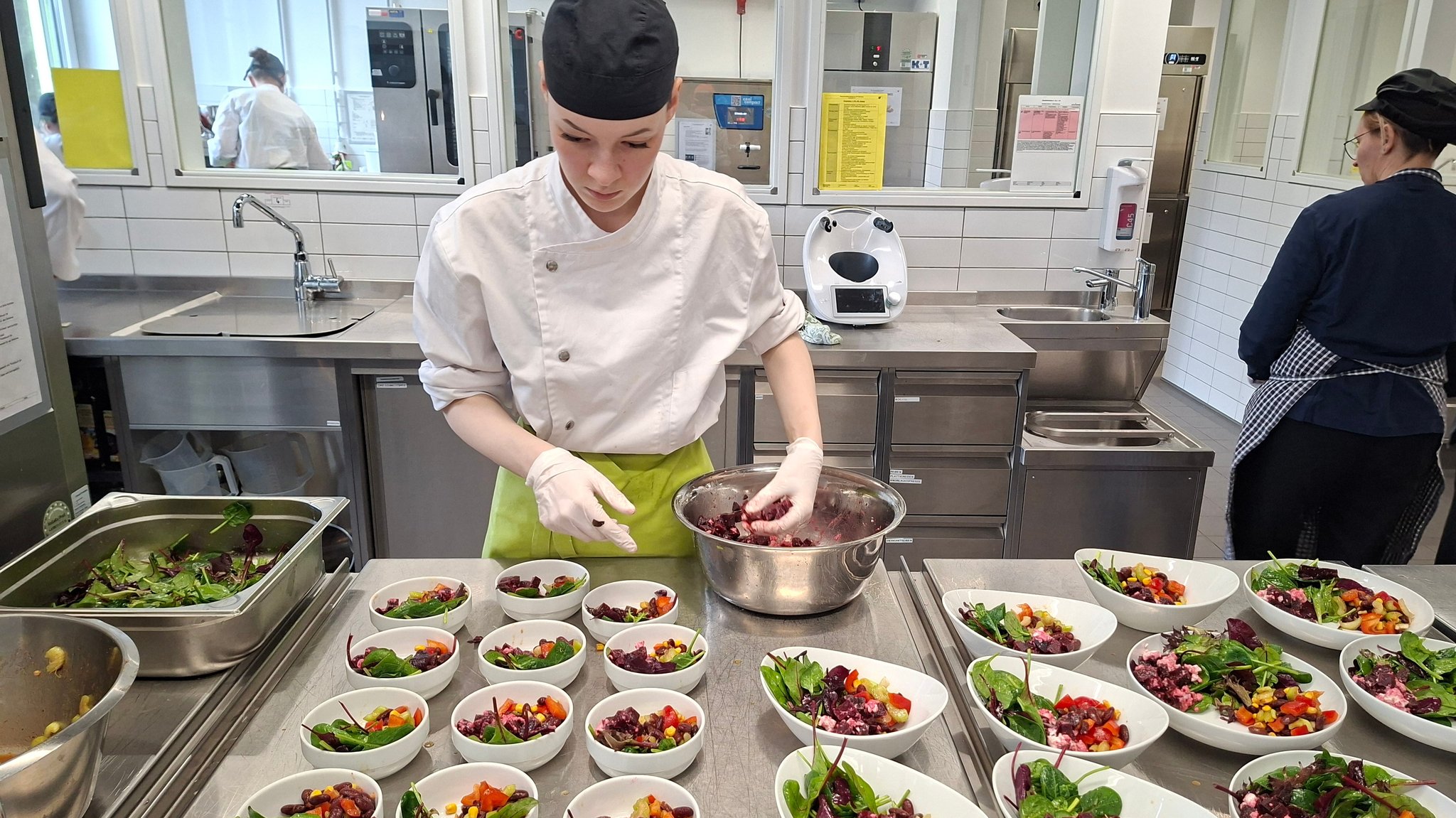 Eine Studentin bereitet in einer Schulküche Salatteller vor. Vor ihr stehen mehrere Schälchen mit verschiedenen Salaten und Gemüse. 