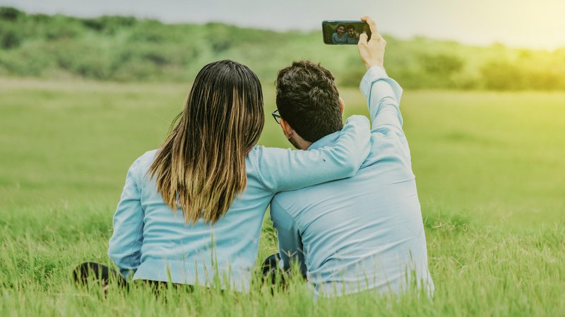 Junges glückliches Paar nimmt ein Selfie im Feld sitzend auf | Bild: picture alliance / imageBROKER | Isai Hernandez Junges glückliches Paar nimmt ein Selfie im Feld sitzend auf