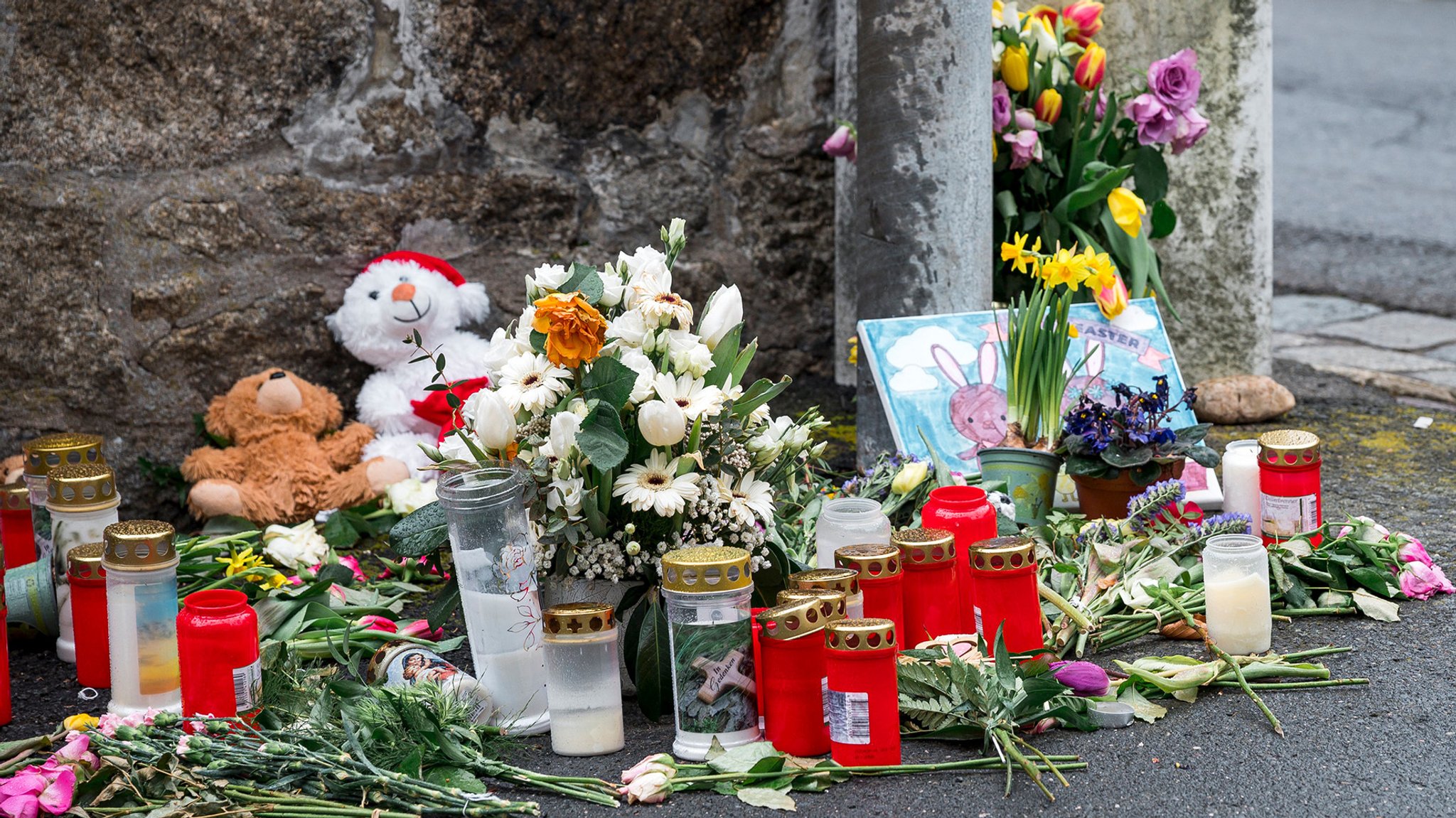 Grabkerzen, Kuscheltiere und Blumen liegen auf dem Boden an einer Mauer. Auch ein selbstgezeichnetes Bild eines Kindes ist zu sehen. 