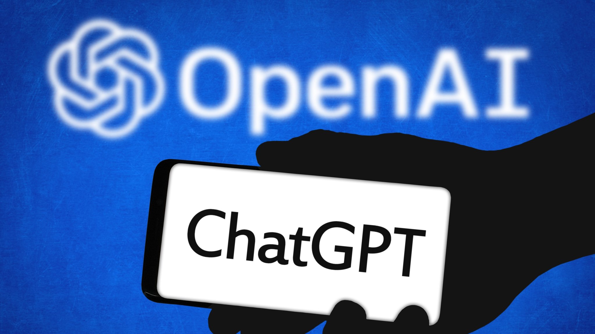 (Symbolbild) Die Silhouette einer Hand hält ein Smartphone mit der Aufschrift "ChatGPT". Im Hintergrund leuchtet das OpenAI-Logo auf einem blauen Hintergrund.