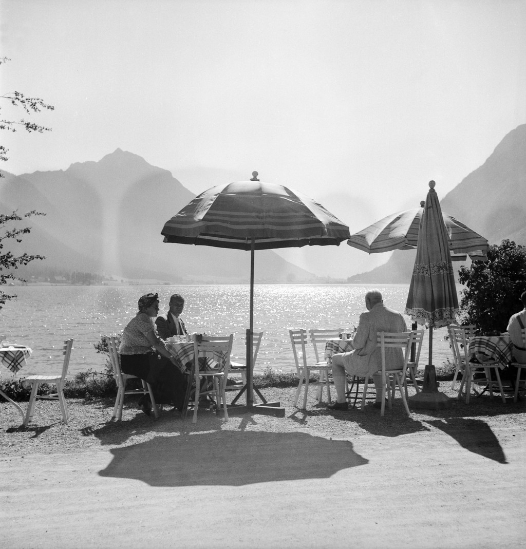 Schwarzweißfotografie einiger Cafétische unter Sonnenschirmen an einem See mit Bergblick