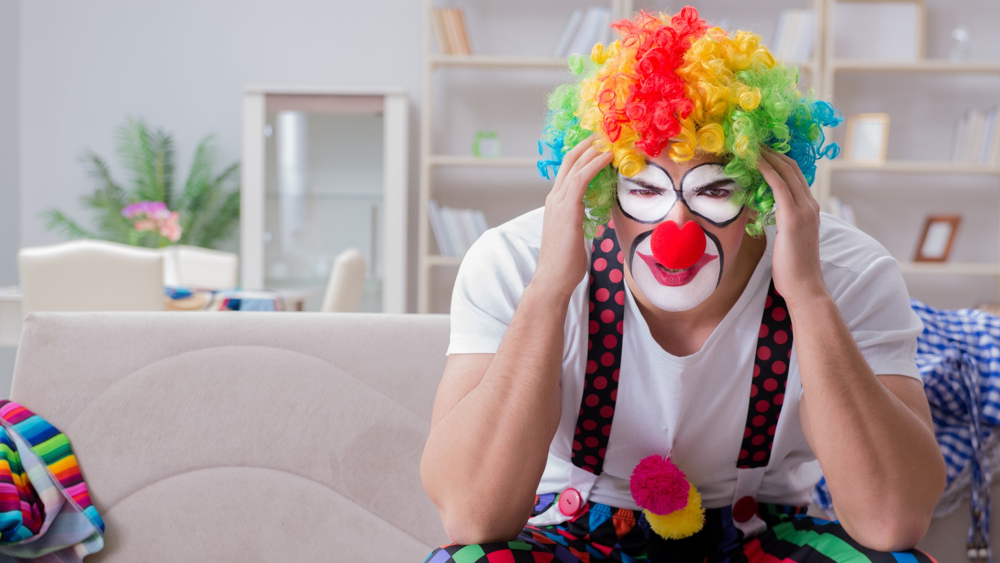 Clown sitzt auf einem Sofa und hält sich schmerzverzerrt den Kopf