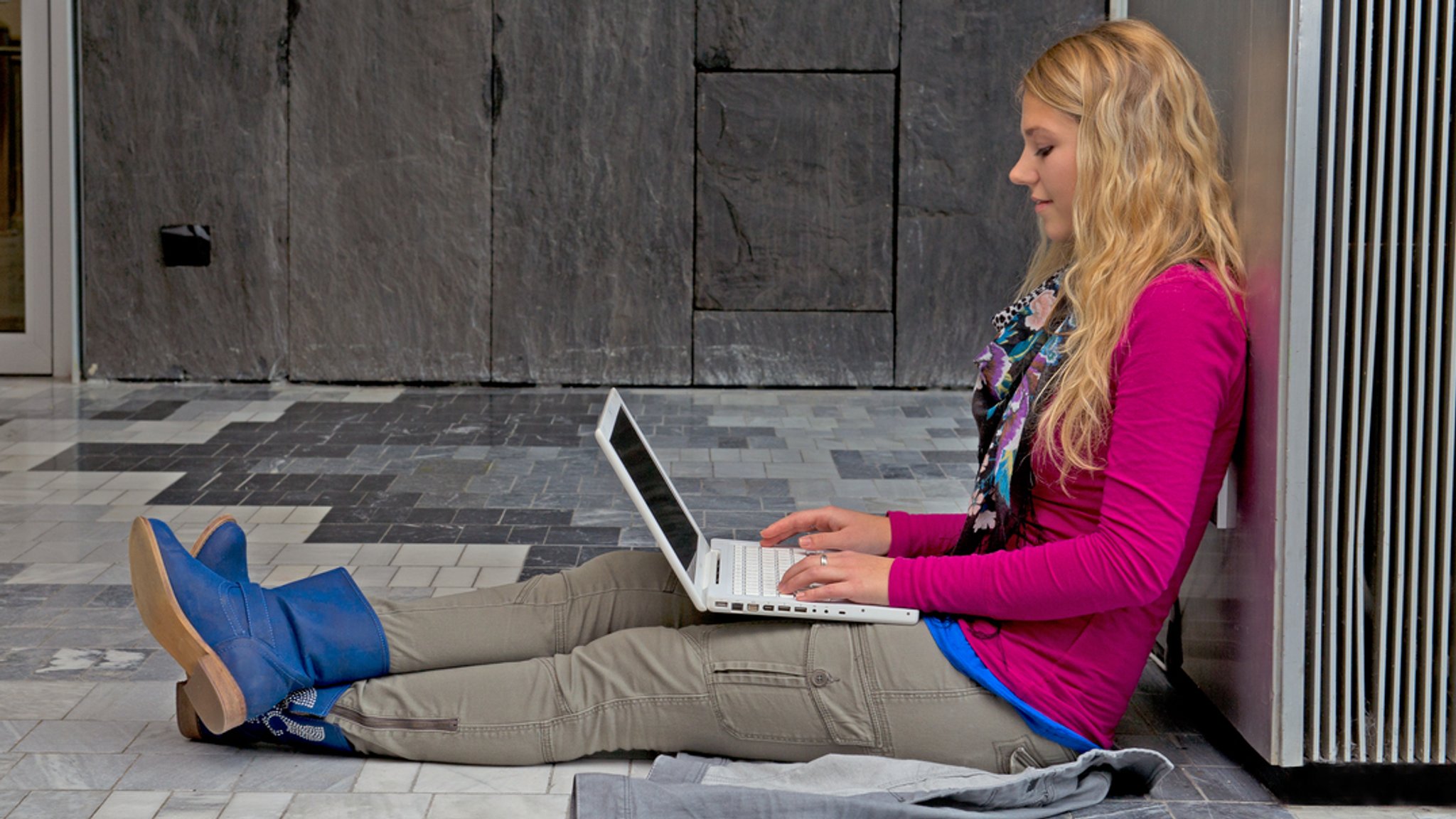 Eine Frau sitzt auf dem Boden mit einem Laptop in der Hand und tippt etwas.