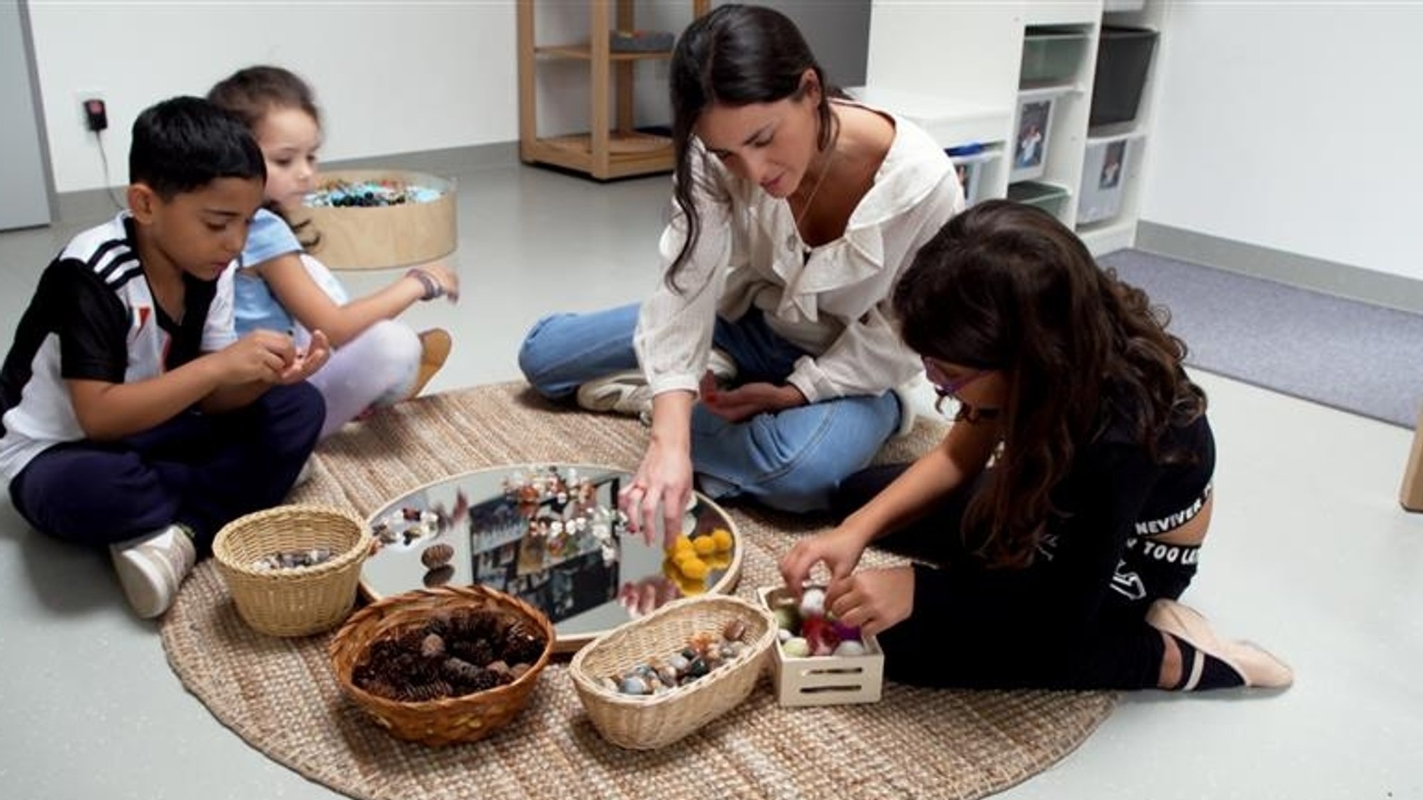 Drei Kinder sitzen auf dem Boden und legen gemeinsam mit einer Erzieherin Steine und Perlen auf einen Spiegel. 