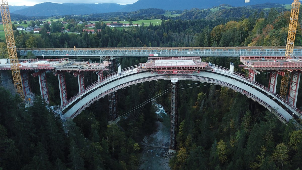 Die Echelsbacher Brücke, eine gebogene Brücke tief über einer bewaldeten Schlucht, durch die die Ammer fließt. Im Hintergrund ein Dorf und Berge.