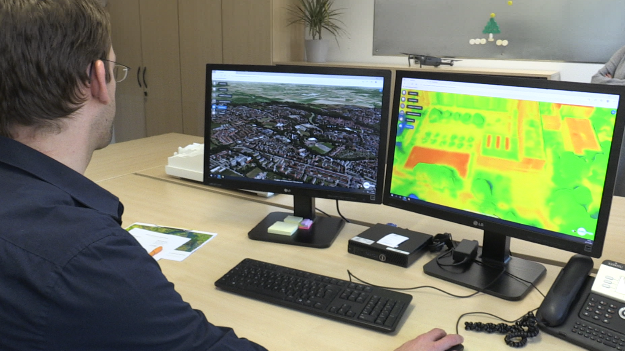 Der Landsberger Stadtplaner Daniel Broschart sitzt vor zwei Monitoren, einer zeigt den Digitalen Zwilling von Landsberg von oben, das andere zeigt die Wärmebildansicht mit roten, gelben und grünen Flächen.