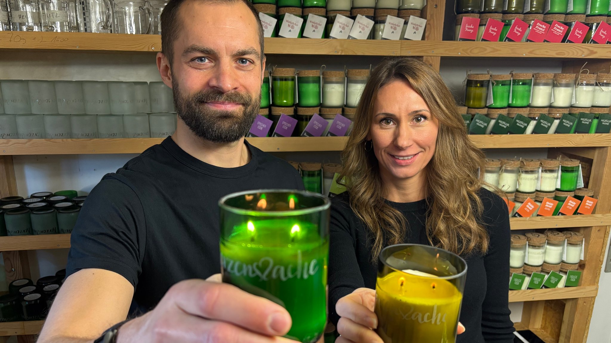 Ein Mann und eine Frau halten brennende Kerzen jeweils in einem Glas in der Hand. Eine ist grün, die andere gelb.