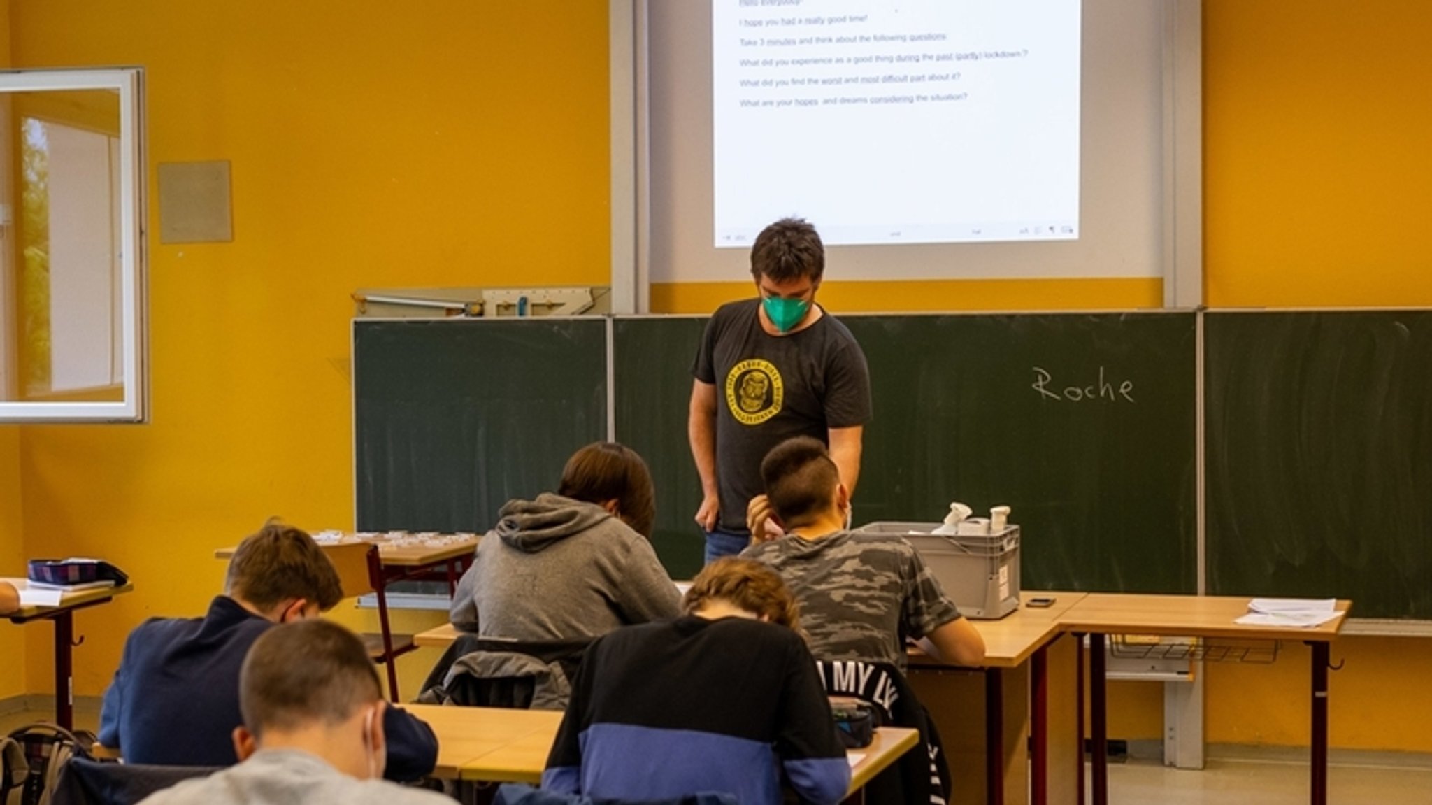 Schülerinnen und Schüler sitzen in ihrer Klasse, während ein Lehrer neben geöffneten Fenstern vor der Tafel steht. 