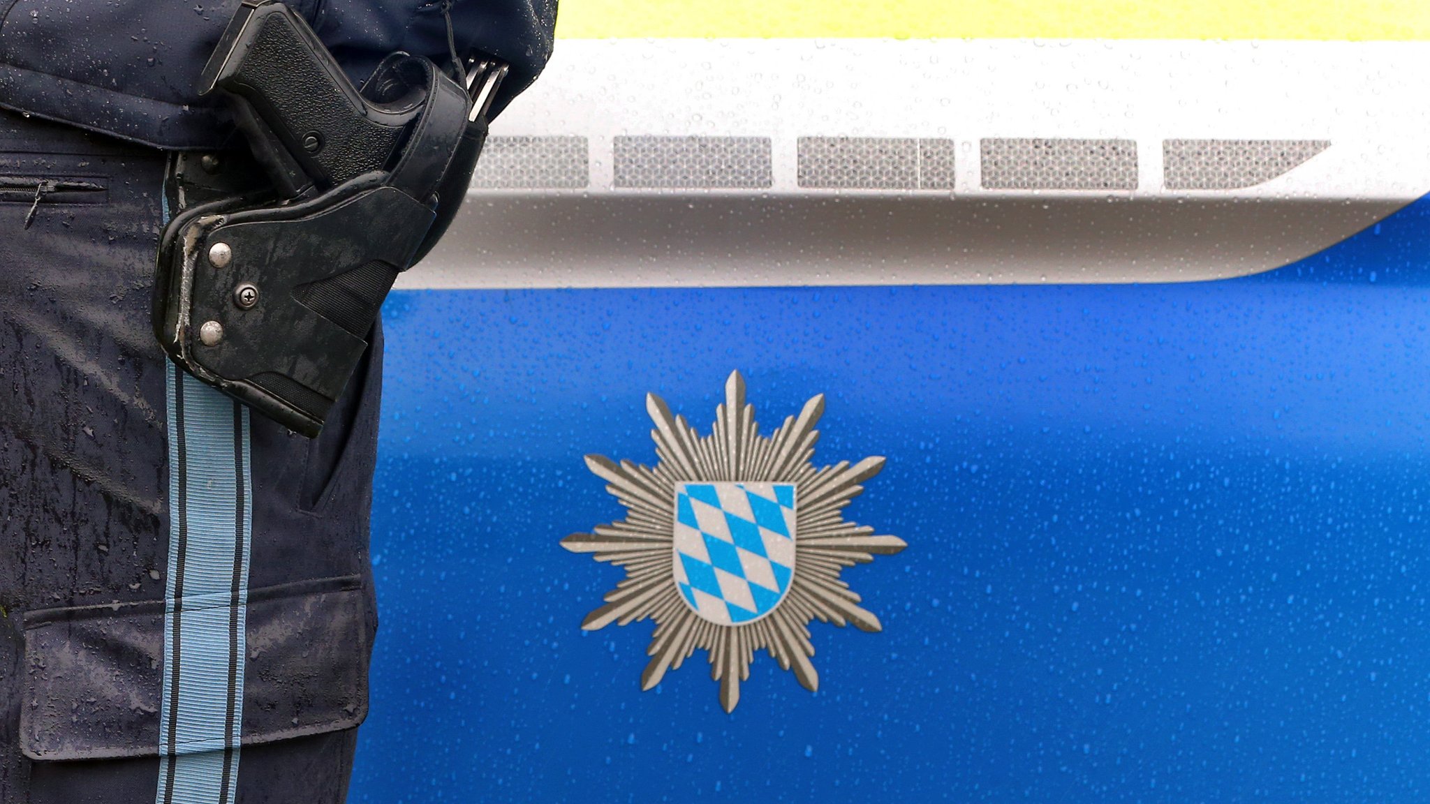 Ein Polizist mit Handschellen und einer Pistole am Gürtel steht vor einem Streifenwagen (Archiv- und Symbolbild)