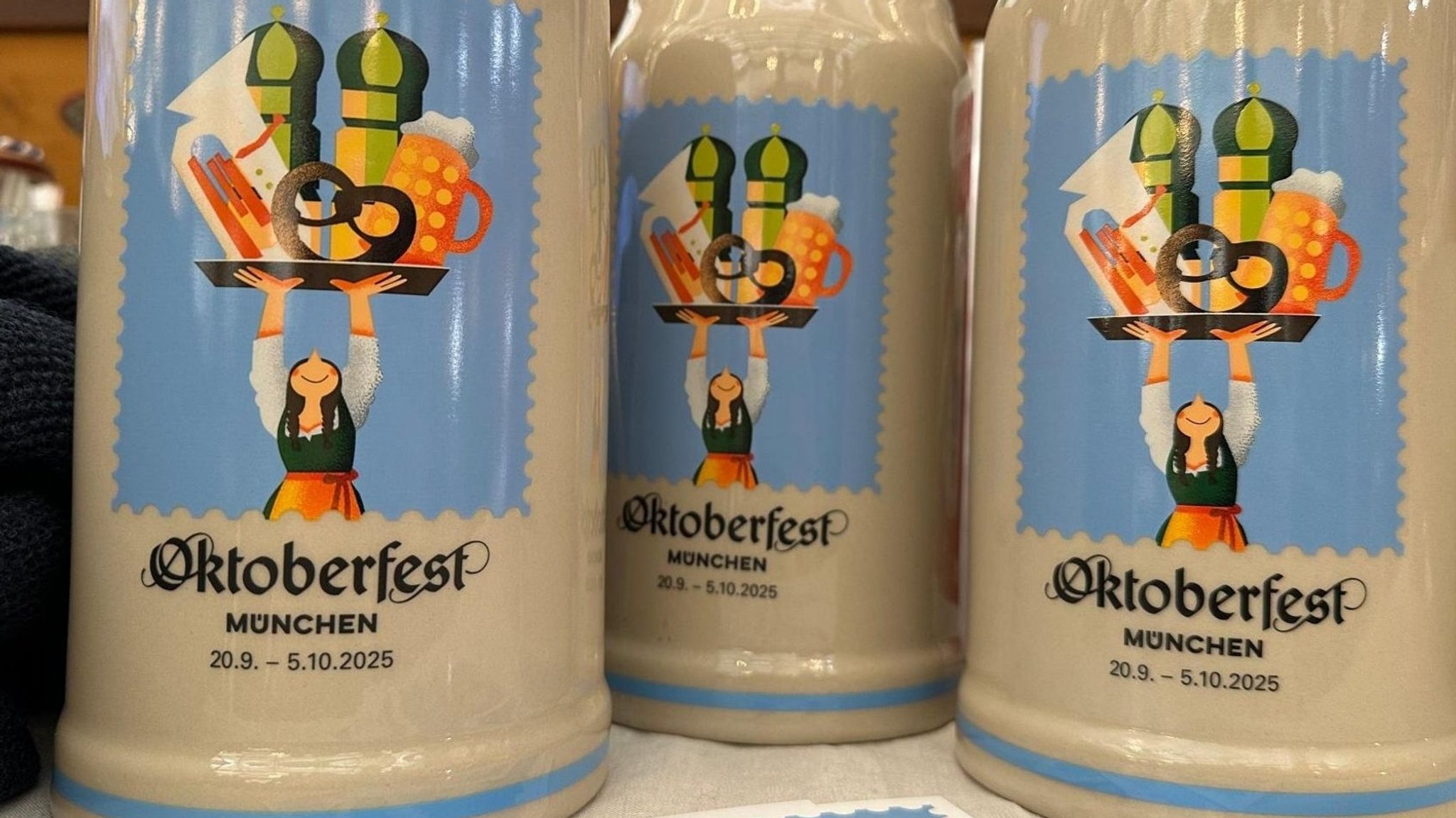 Drei Bierkrüge mit Logo vom Oktoberfest 2025 stehen auf einem Tisch. 