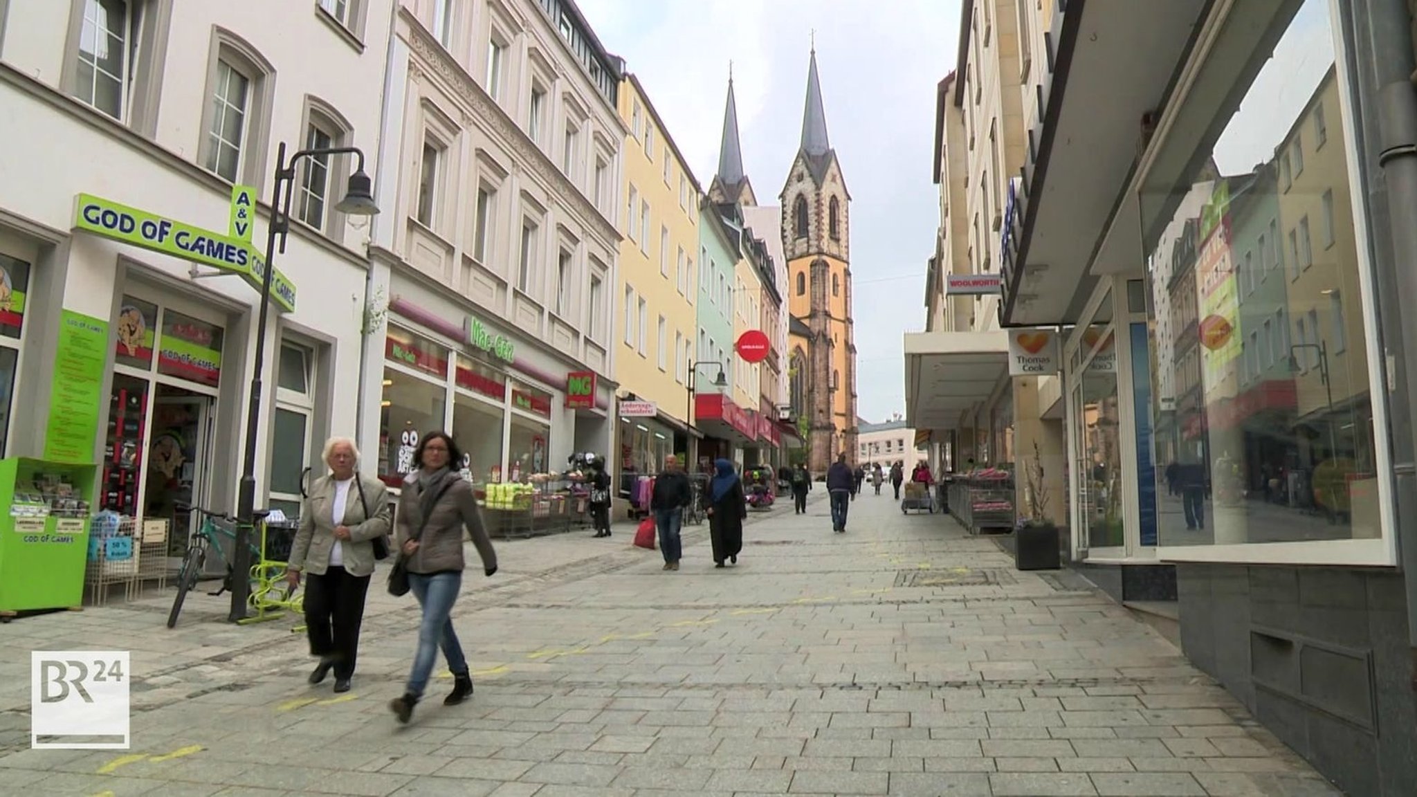 Passanten gehen durch die Fußgängerzone in Hof, im Hintergrund die Kirche mit ihren zwei Türmen. 