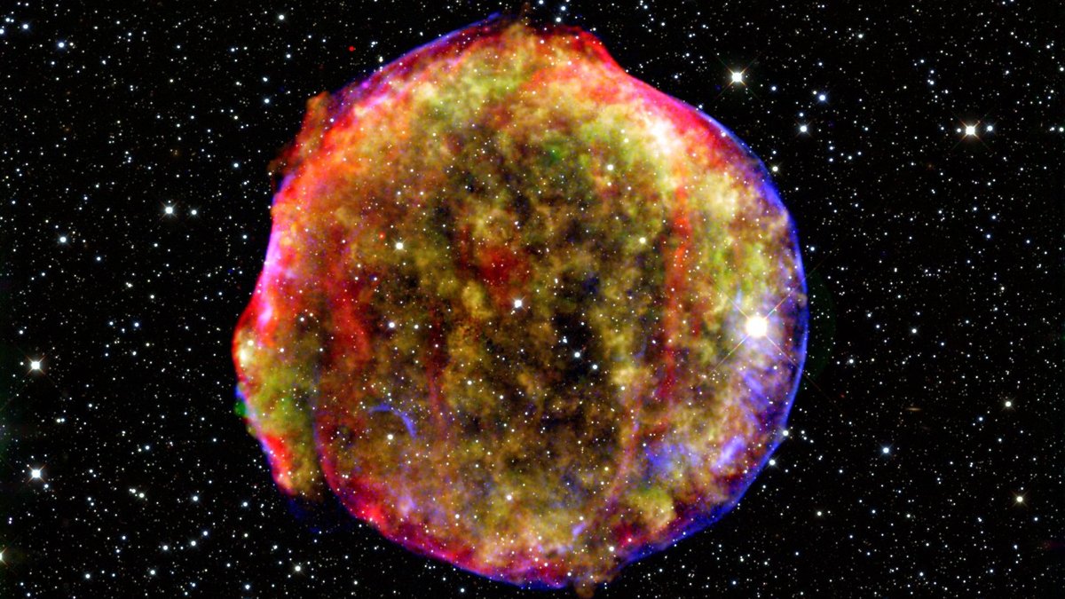 Vor einem dunklen Hintergrund voller Sterne befindet sich eine etwa kreisförmige, bunte Wolke, die in verschiedenen Farben eingefärbt ist. Diese Wolke ist der Überrest einer Supernova vom Typ 1a.