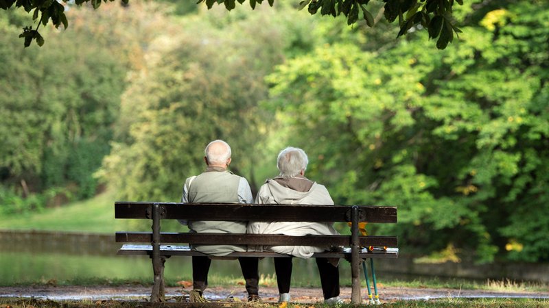 Ein Rentnerpaar sitzt in einem Park auf der Bank vor einem Teich. Im Hintergrund grüne Bäume in Unschärfe. (Symbolbild)