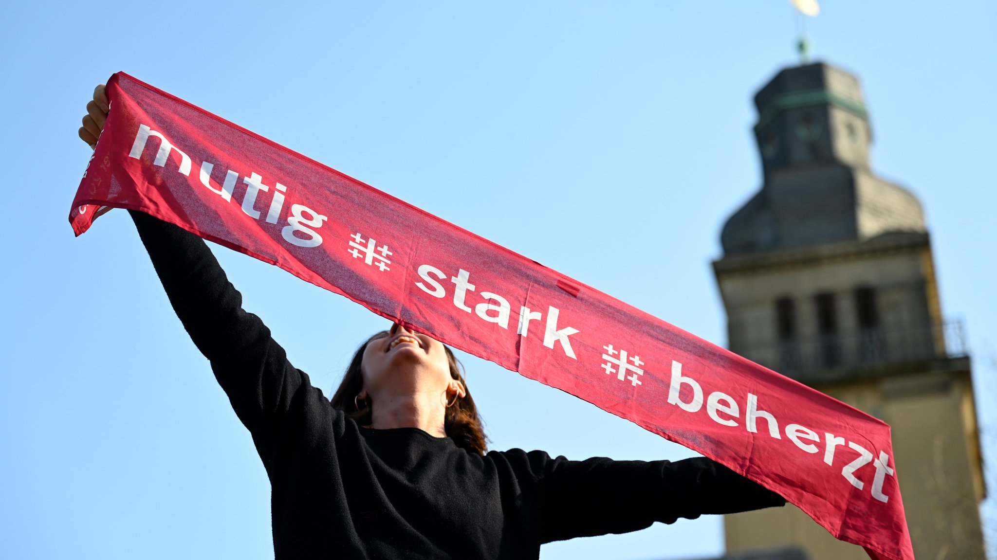 Eine Frau hält einen Schal in der Luft. Darauf das Motto des Kirchentags: "mutig, stark, beherzt". im Hintergund verschwommen ist ein Kirchturm. | Bild: picture alliance / epd-bild | Heike Lyding Eine Frau hält einen Schal in der Luft. Darauf das Motto des Kirchentags: "mutig, stark, beherzt". im Hintergund verschwommen ist ein Kirchturm.