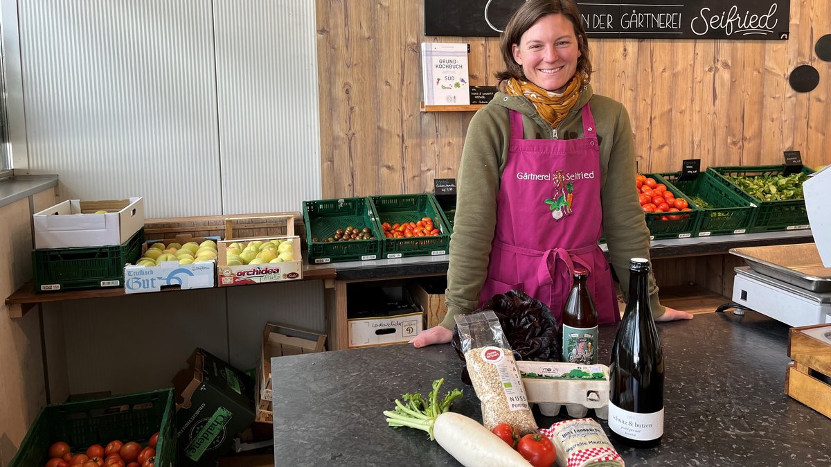 Hofladenbesitzerin Verena Seifried steht an der Kasse im Hofladen, hinter ihr Gemüse, vor ihr andere regionale Produkte wie Apfelschaumwein oder Maultaschen.