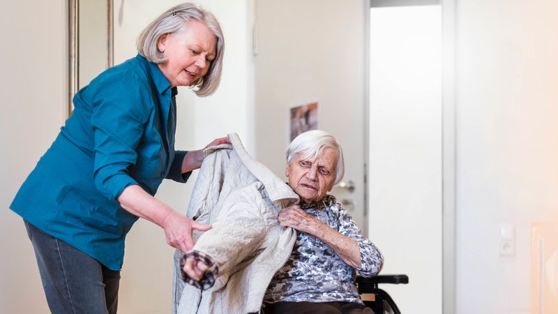 (Symbolbild) Eine Pflegerin hilft einer älteren Dame im Rollstuhl dabei, eine Jacke anzuziehen. | Bild: pa/dpa/Daniel Ingold (Symbolbild) Eine Pflegerin hilft einer älteren Dame im Rollstuhl dabei, eine Jacke anzuziehen.