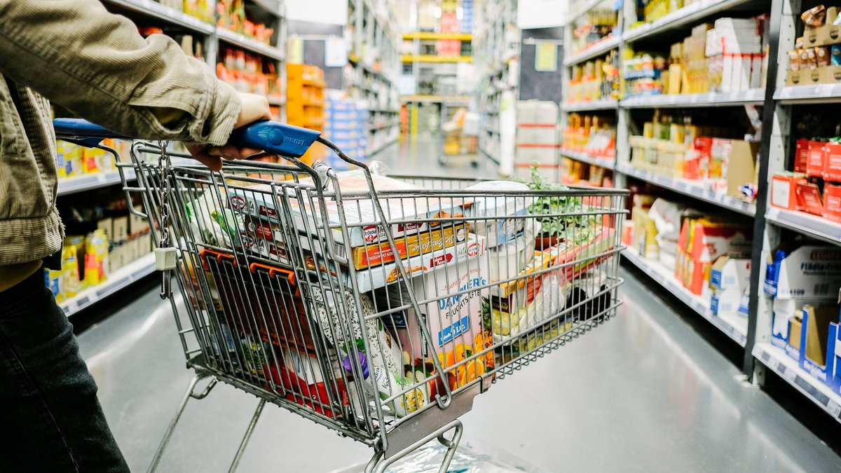 Eine Person schiebt einen vollen Einkaufswagen durch einen Supermarkt.