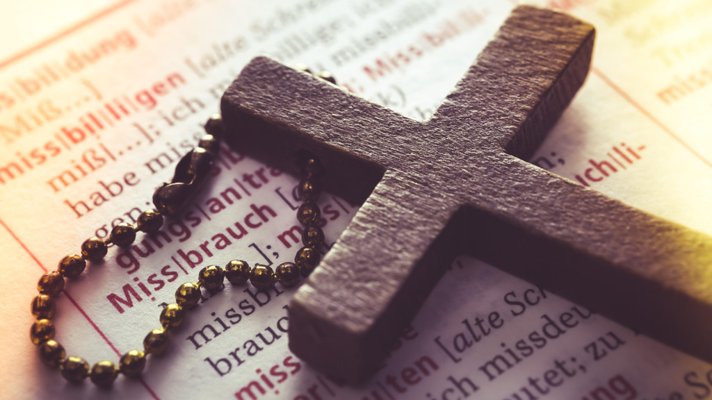 Ein Kreuz an einem Rosenkranz vor der aufgeschlagenen Seite eines Wörterbuches, wo das Wort "Missbrauch" zu lesen ist.