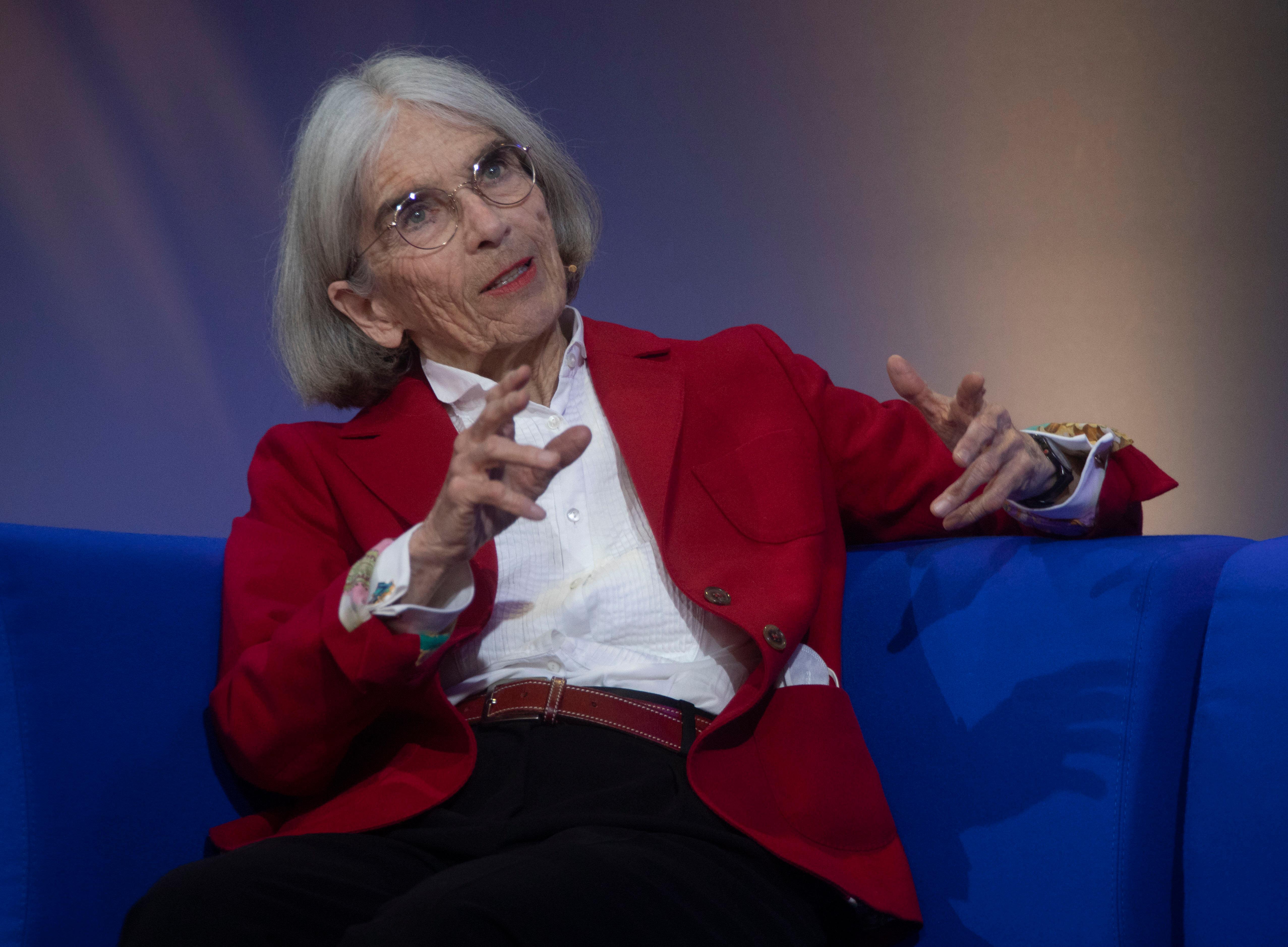 Donna Leon sitzt in weißer Bluse und rotem Blazer auf einem blauen Sofa. (Archivbild) Sie erhält im November den Ehrenpreis des Bayerischen Buchpreises. 
