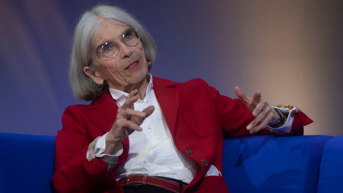 Donna Leon sitzt in weißer Bluse und rotem Blazer auf einem blauen Sofa. (Archivbild) Sie erhält im November den Ehrenpreis des Bayerischen Buchpreises. 