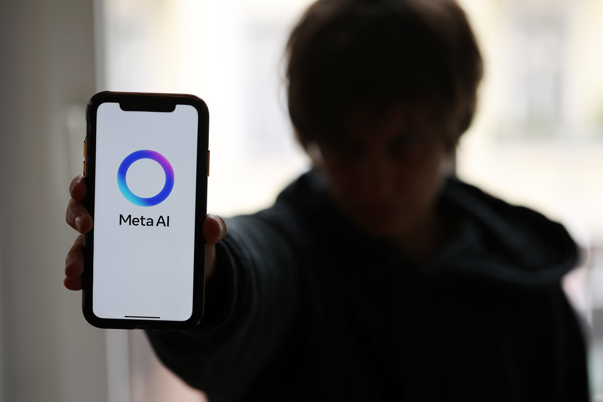 Symbolfoto. Eine junge Person im Kapuzenpullover hält ein Smartphone mit dem Logo von Meta AI in die Kamera.