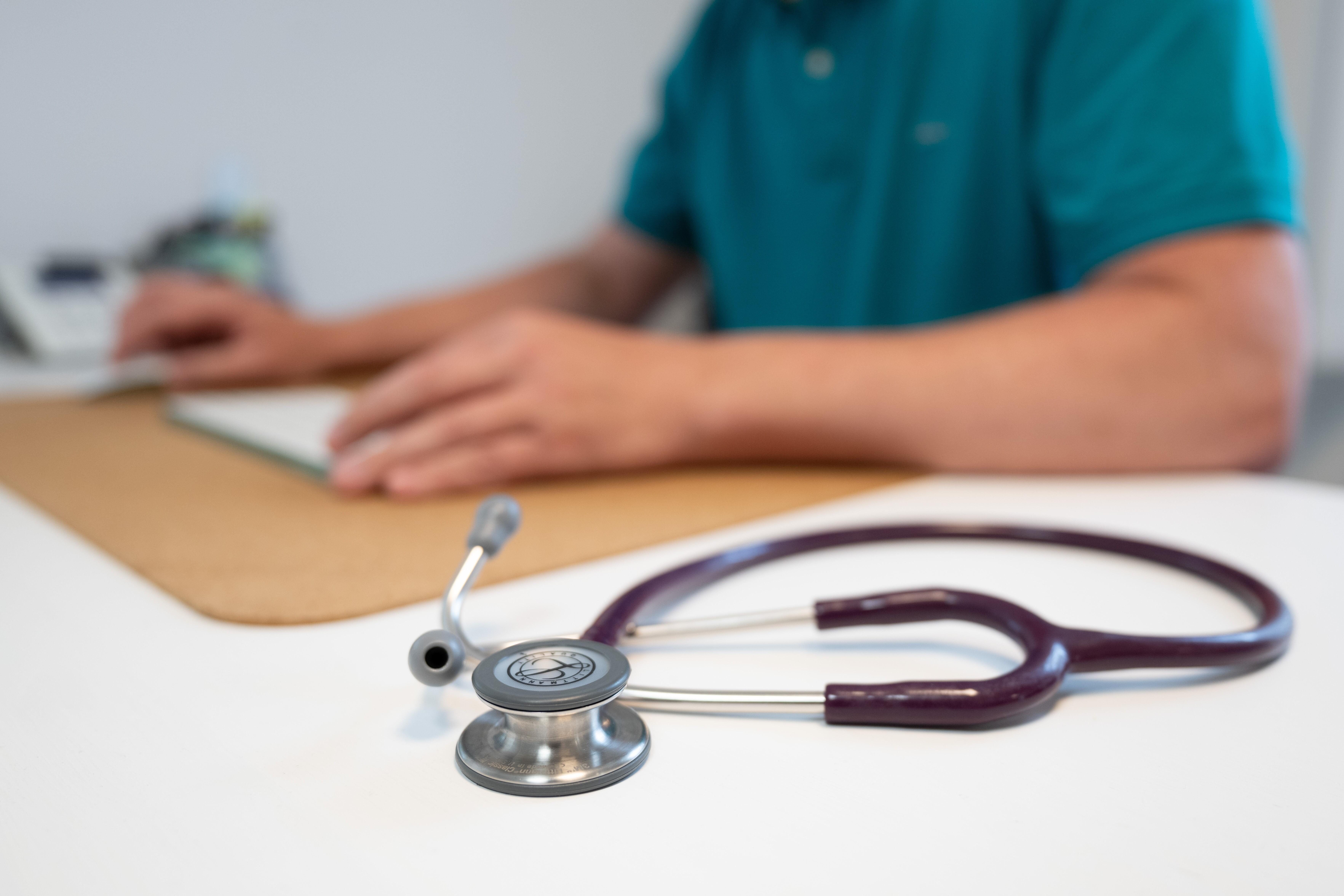 Ein Hausarzt sitzt in einem Sprechzimmer seiner Hausarztpraxis an einem Schreibtisch neben einem Stethoskop und arbeitet am Computer. 